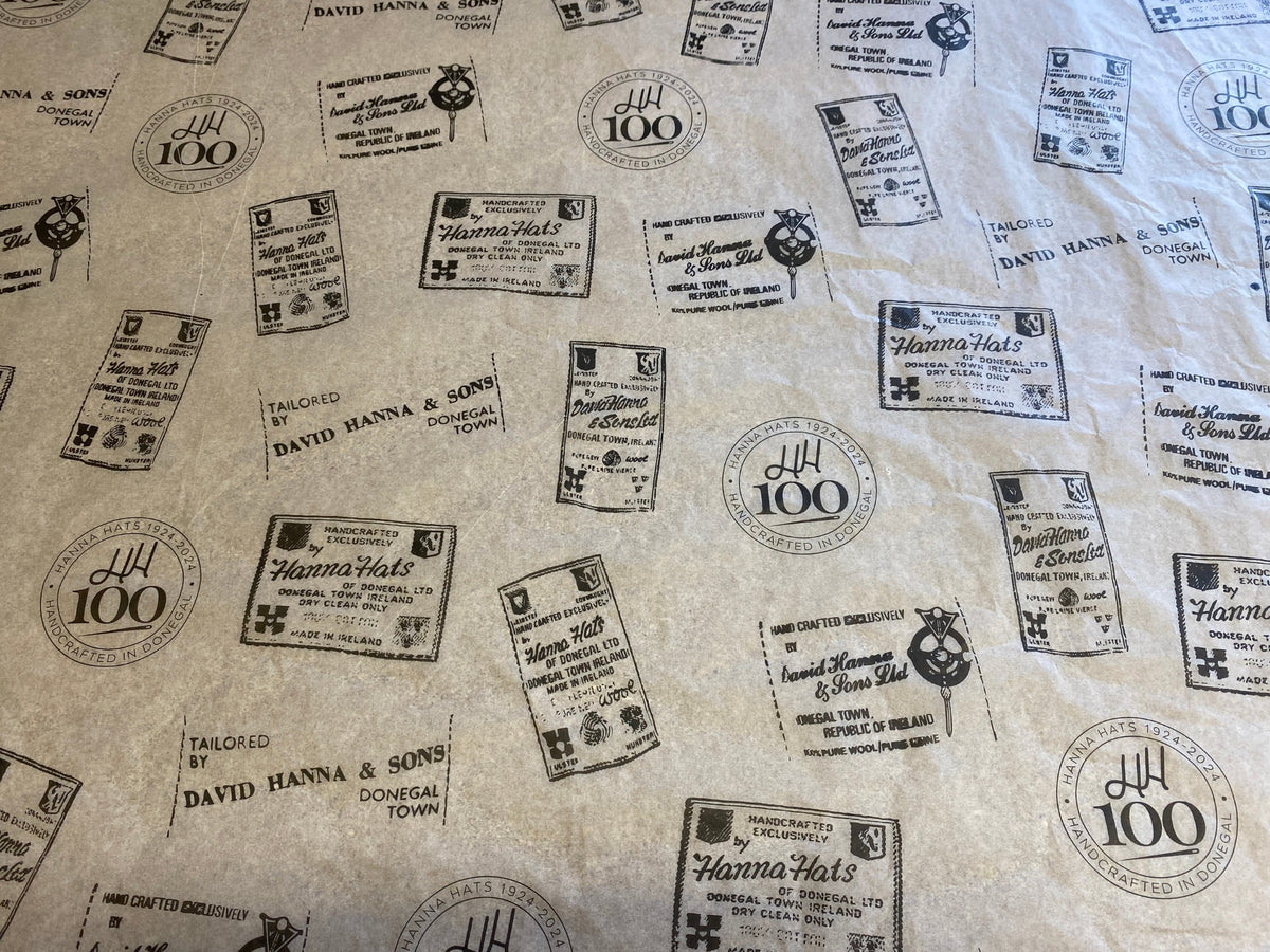 Brown paper printed with various vintage-style black stamps and labels, including “Hanna Hats,” “David Hanna & Sons,” and “Tailored in Donegal Town,” along with coat of arms and anniversary logos.