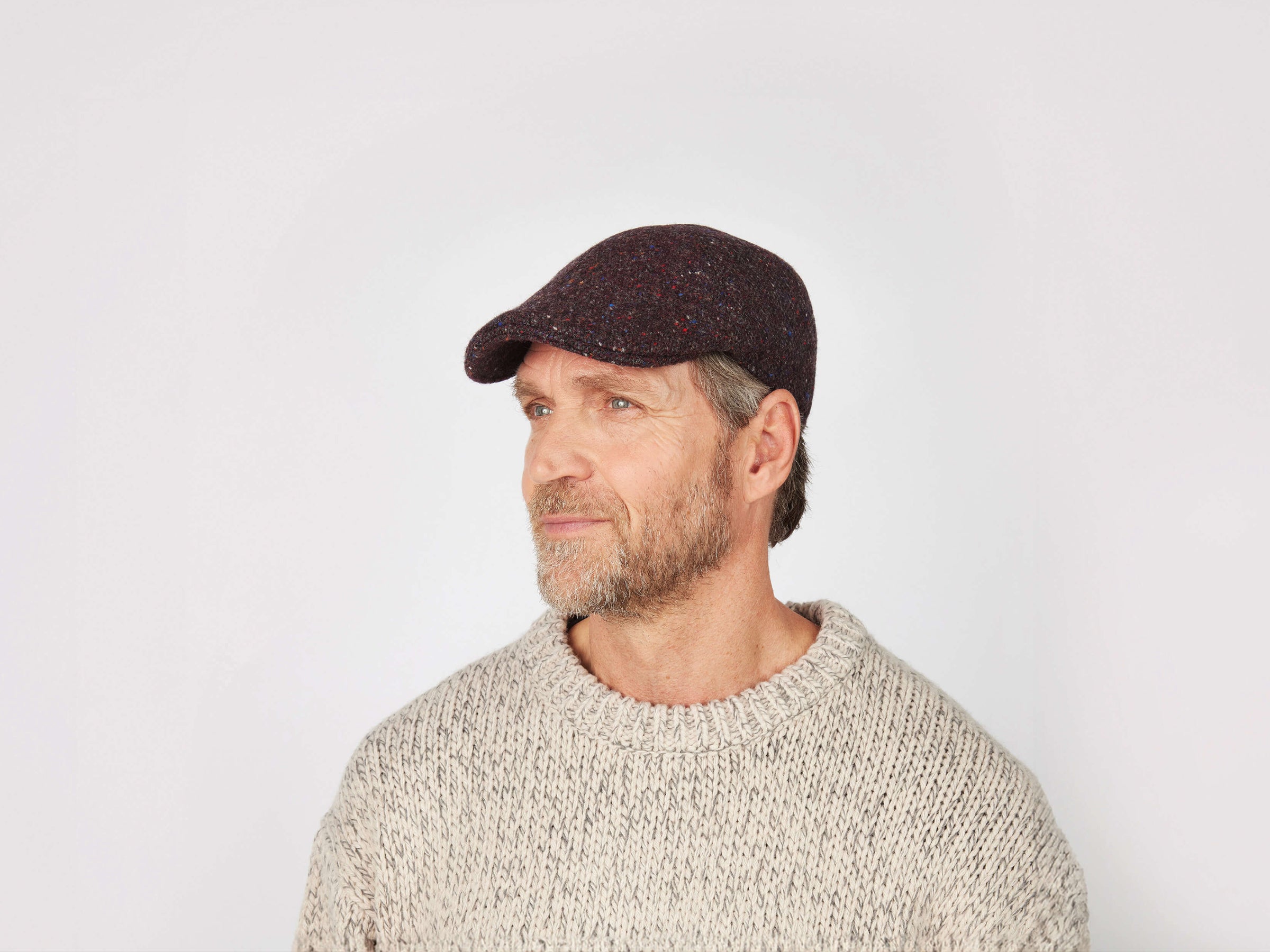A middle-aged bearded man wears a textured beige sweater and the Erin Cap Tweed by Hanna Hats, looking left against a plain white background.