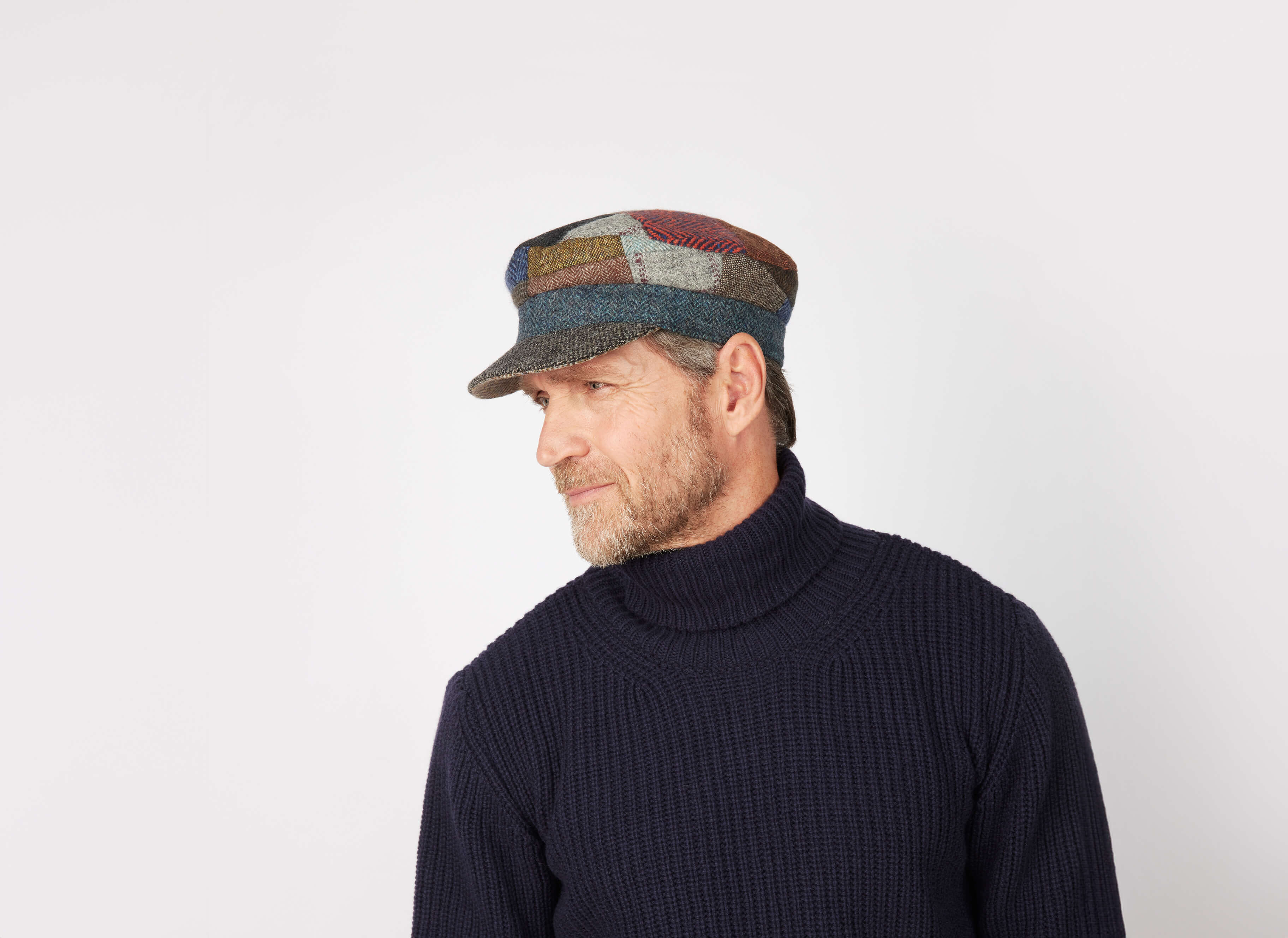 A bearded man in a dark blue turtleneck and a Hanna Hats Skipper Cap Patchwork Tweed looks to his left against a plain white background.