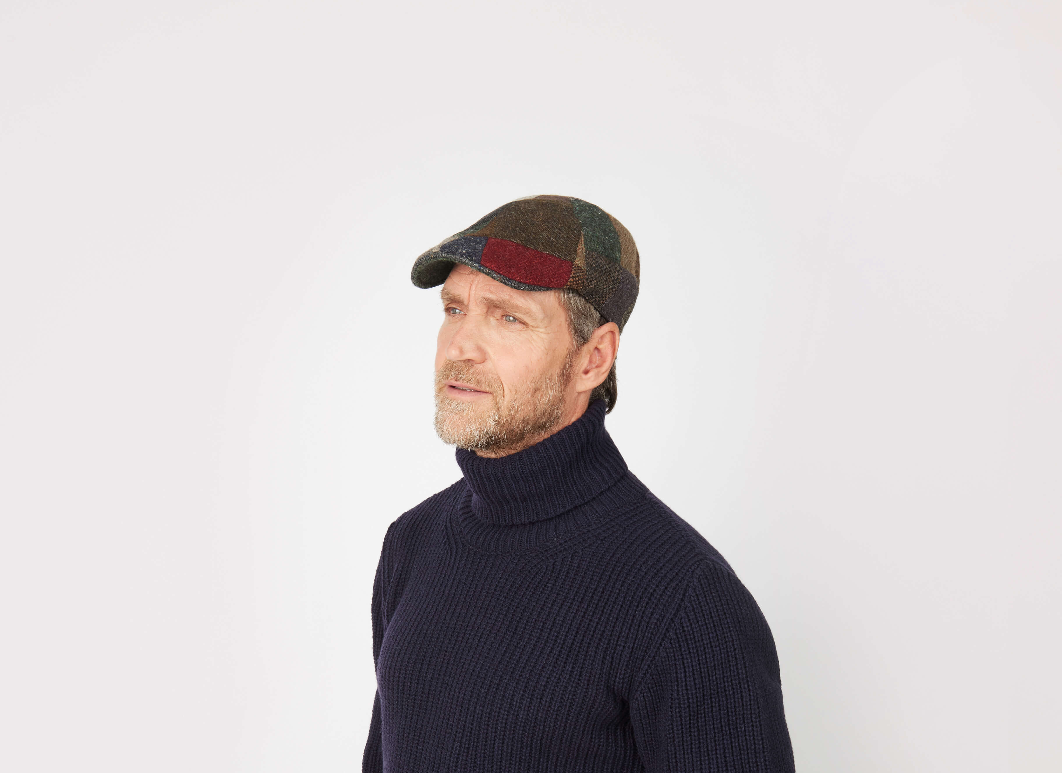 A bearded man in a dark turtleneck and the Hanna Hats Erin Cap Patchwork Tweed gazes slightly to the side against a plain white background.