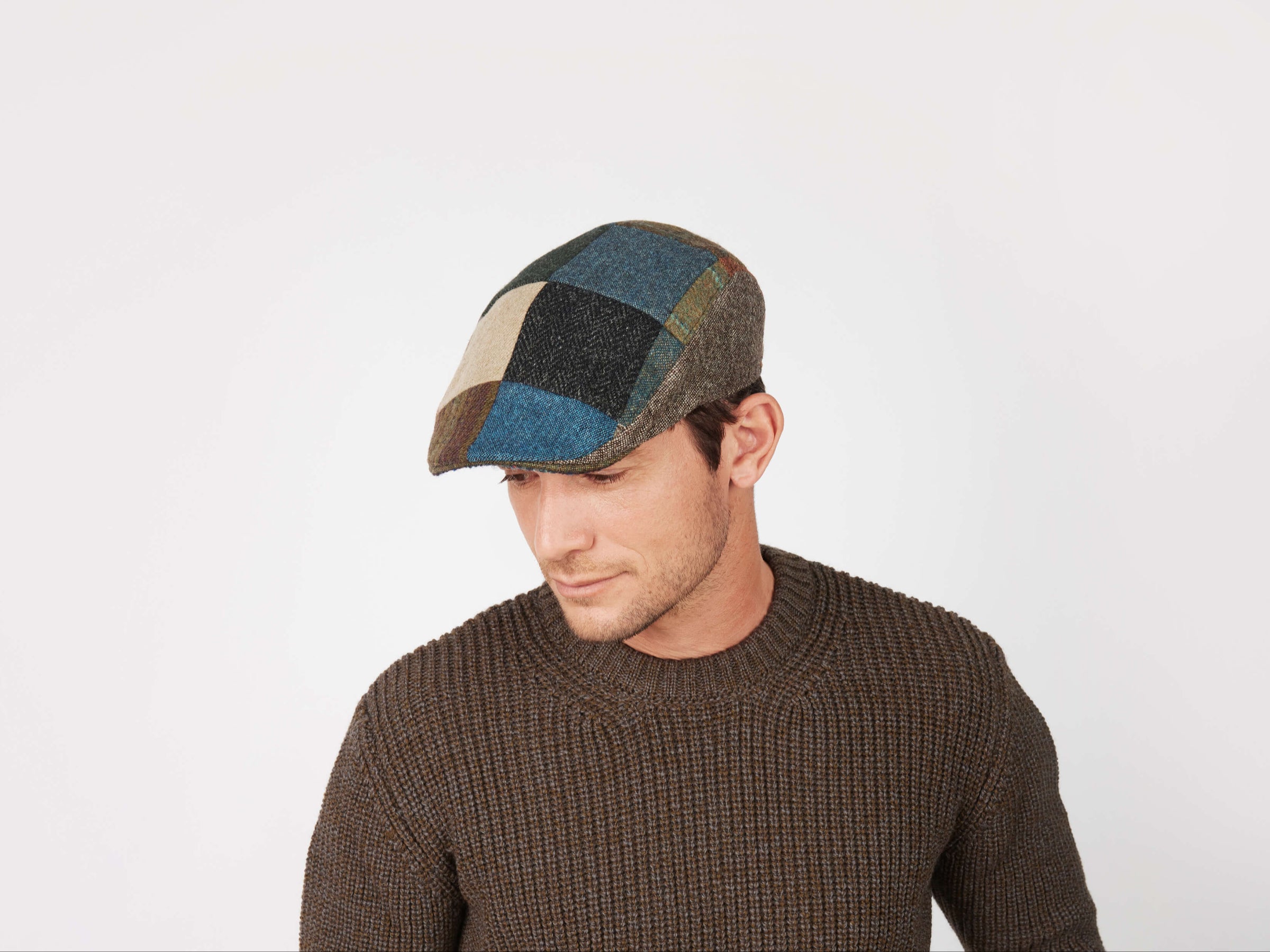 A man in a brown textured sweater and a Hanna Hats Donegal Touring Cap Patchwork Tweed looks down and slightly to his left against a plain white background.