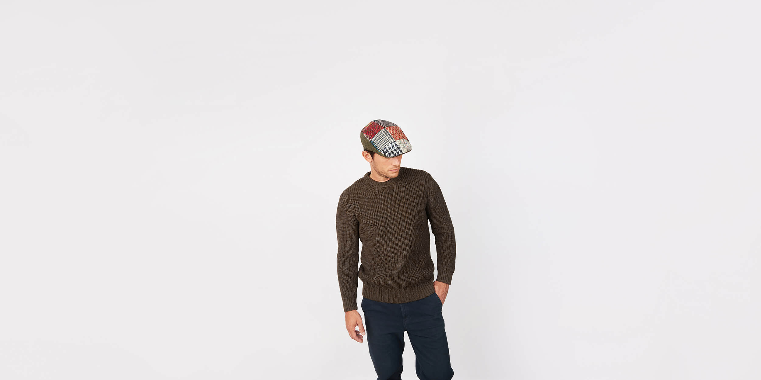 A person wearing a brown sweater, dark pants, and a patchwork flat cap stands against a plain white background, looking downward with one hand in their pocket.
