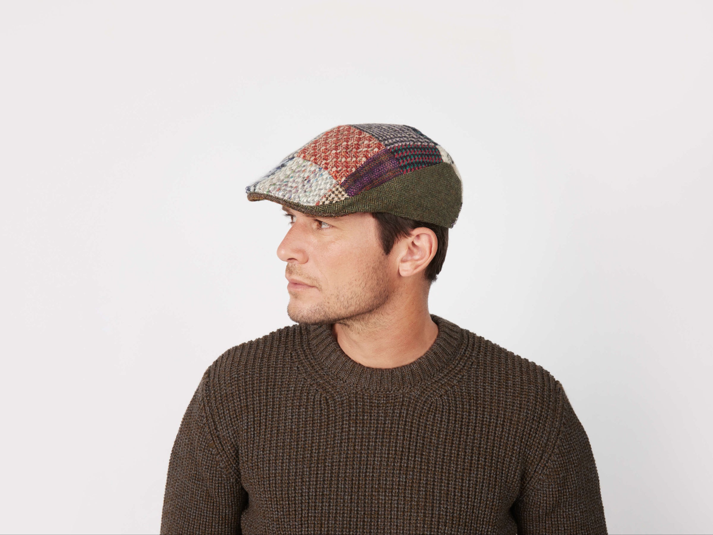A man in a brown knit sweater and the Hanna Hats Donegal Touring Cap Heavy Patchwork Tweed - LIMITED EDITION looks to his left against a plain white background.