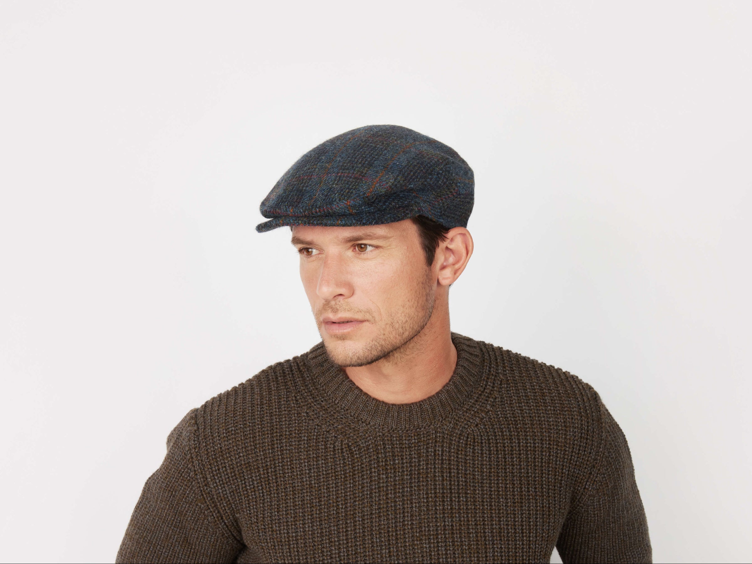 A man wearing a dark knit sweater and the Vintage Cap Tweed by Hanna Hats looks to the side against a plain white background.