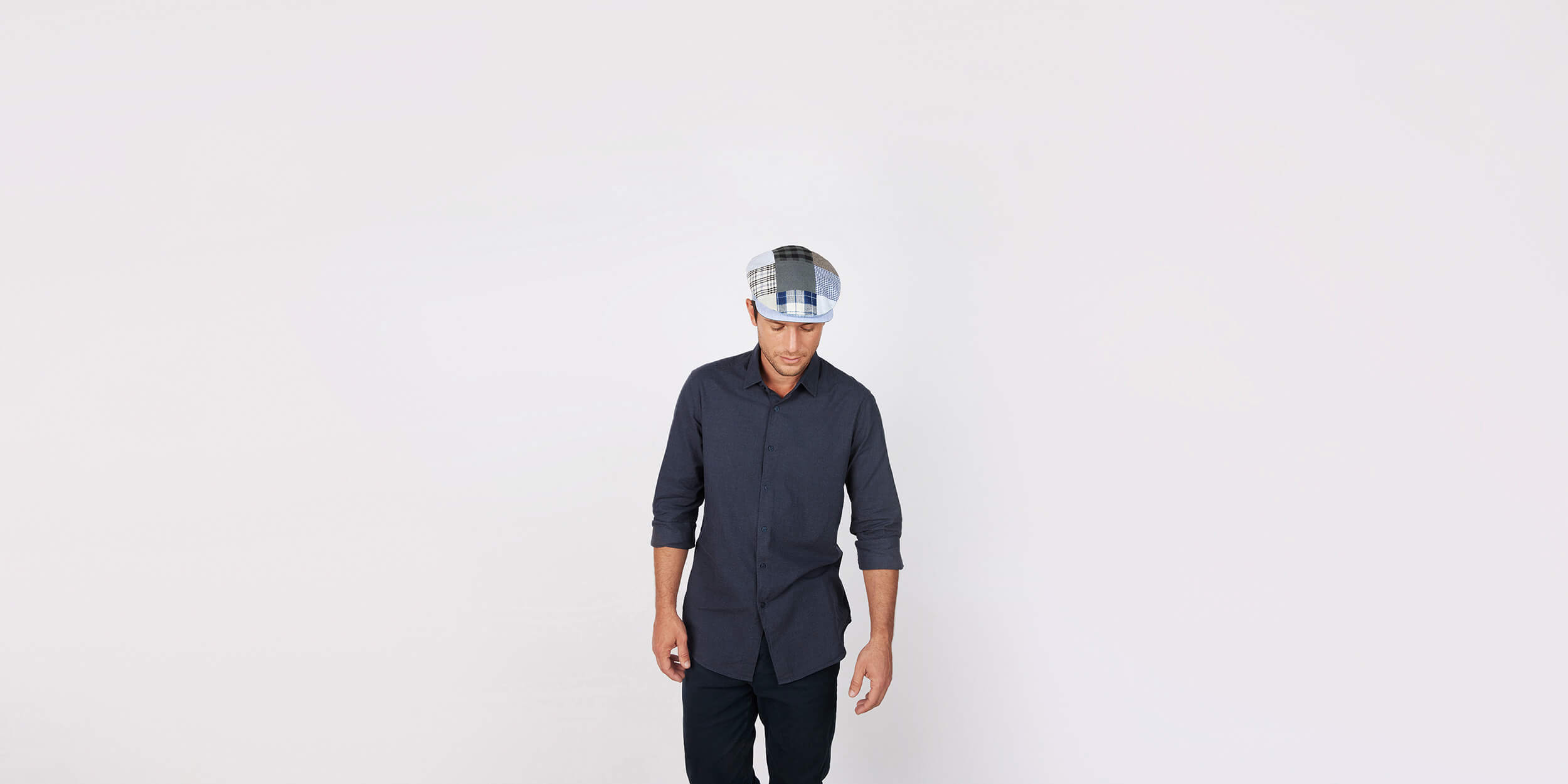 A man wearing a dark button-up shirt, dark pants, and a patterned flat cap looks down with his arms relaxed at his sides, standing against a plain light background.