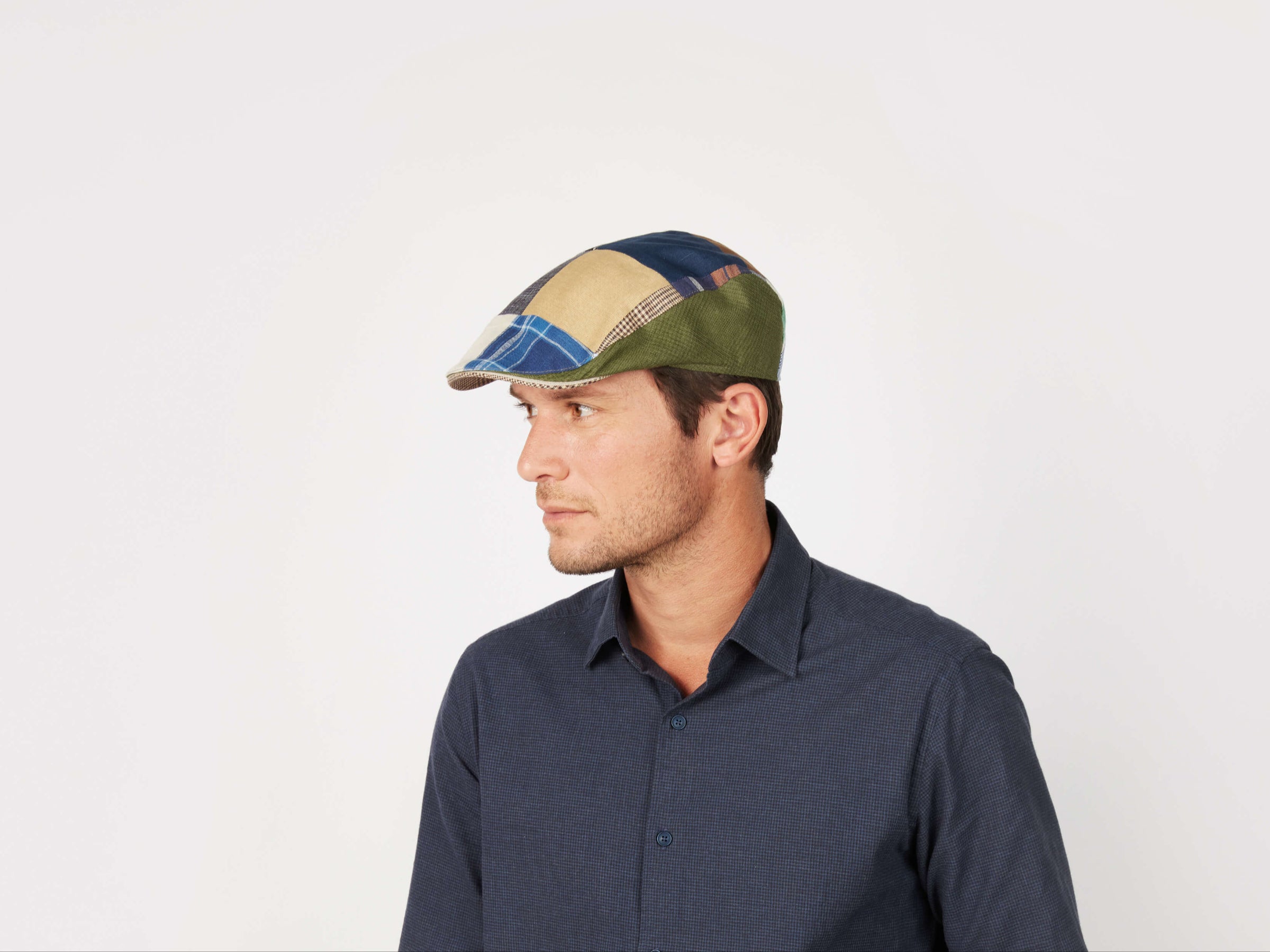 A man in a dark blue button-up shirt and a tailored fit Donegal Touring Cap Patchwork Linen by Hanna Hats looks to the left against a plain white background.