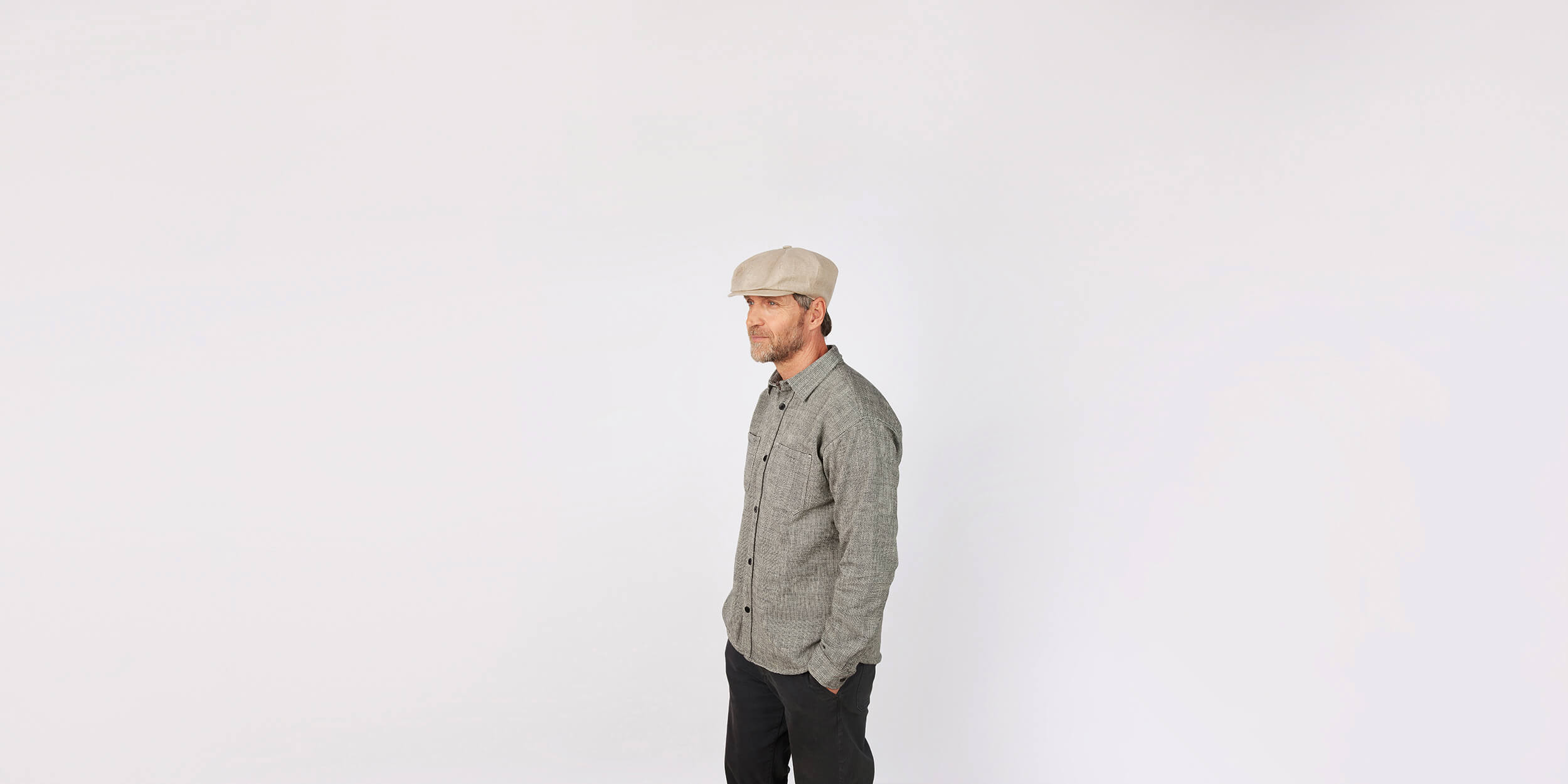 A man wearing a flat cap, grey shirt, and dark pants stands against a plain white background, looking to the left with his hands in his pockets.