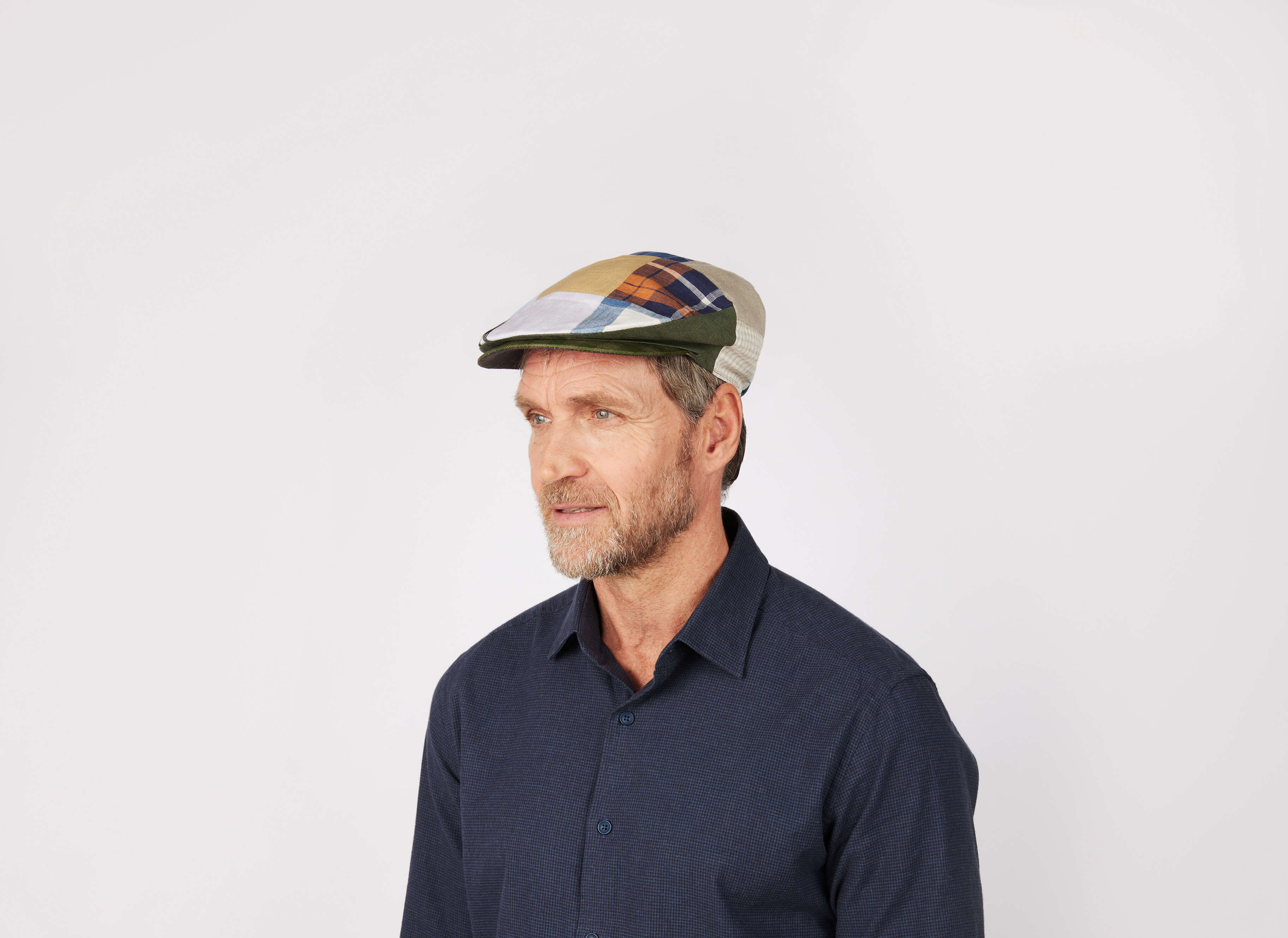 A middle-aged man with a short beard, wearing a dark blue shirt and the Hanna Hats Vintage Cap Patchwork Linen, looks to the side against a plain white background.