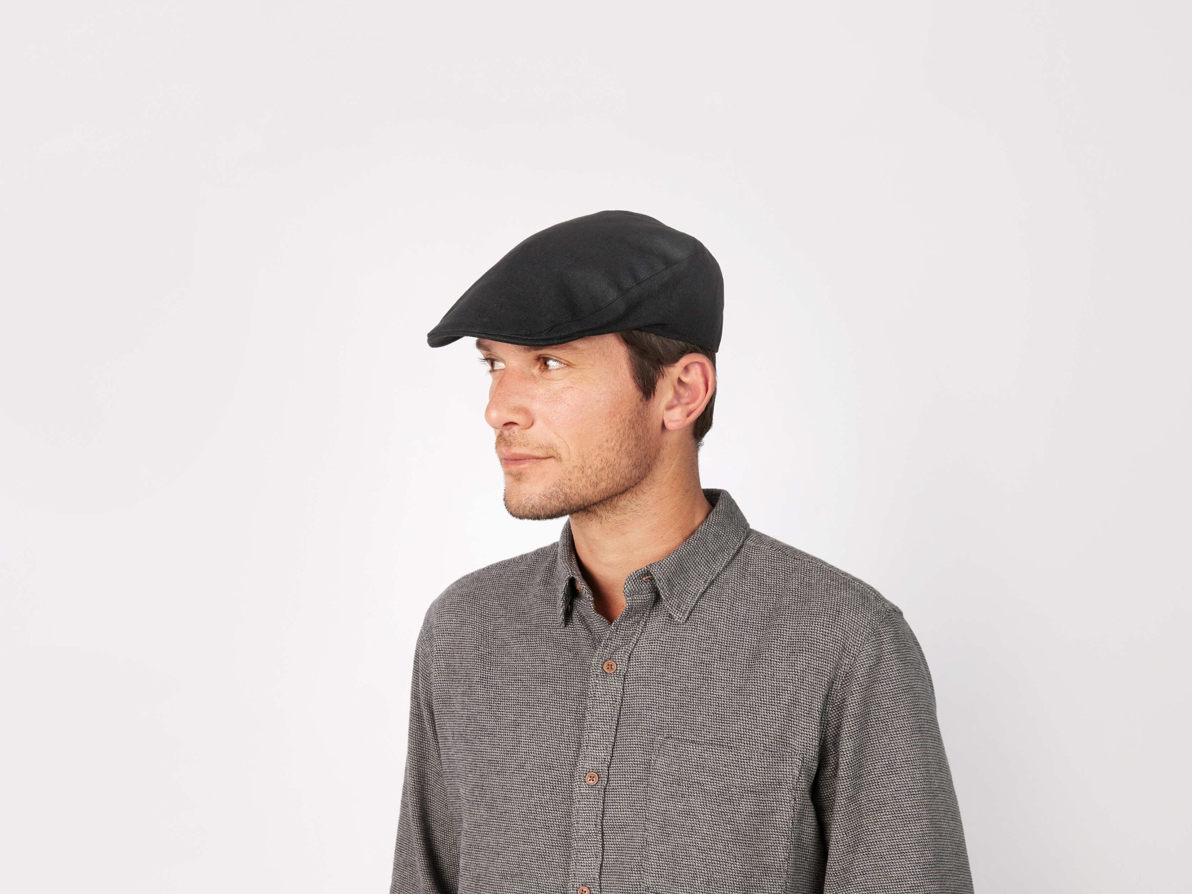 A man wearing a tailored fit Hanna Hats Donegal Touring Cap Linen and a grey button-up shirt looks to his left against a plain white background.