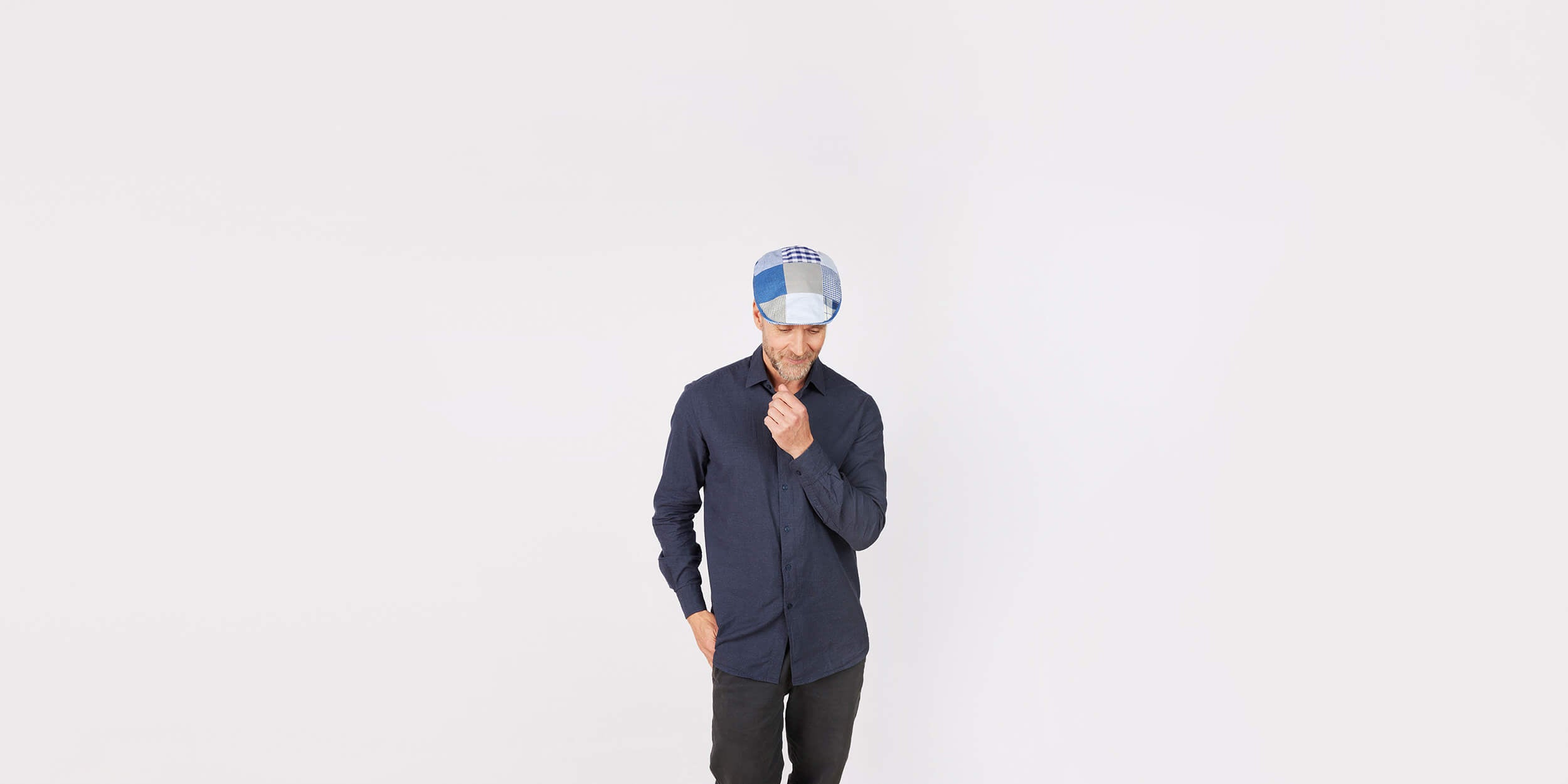 A man wearing a blue shirt and a blue-and-white cap stands against a plain white background, looking down thoughtfully with one hand on his chin and the other in his pocket.