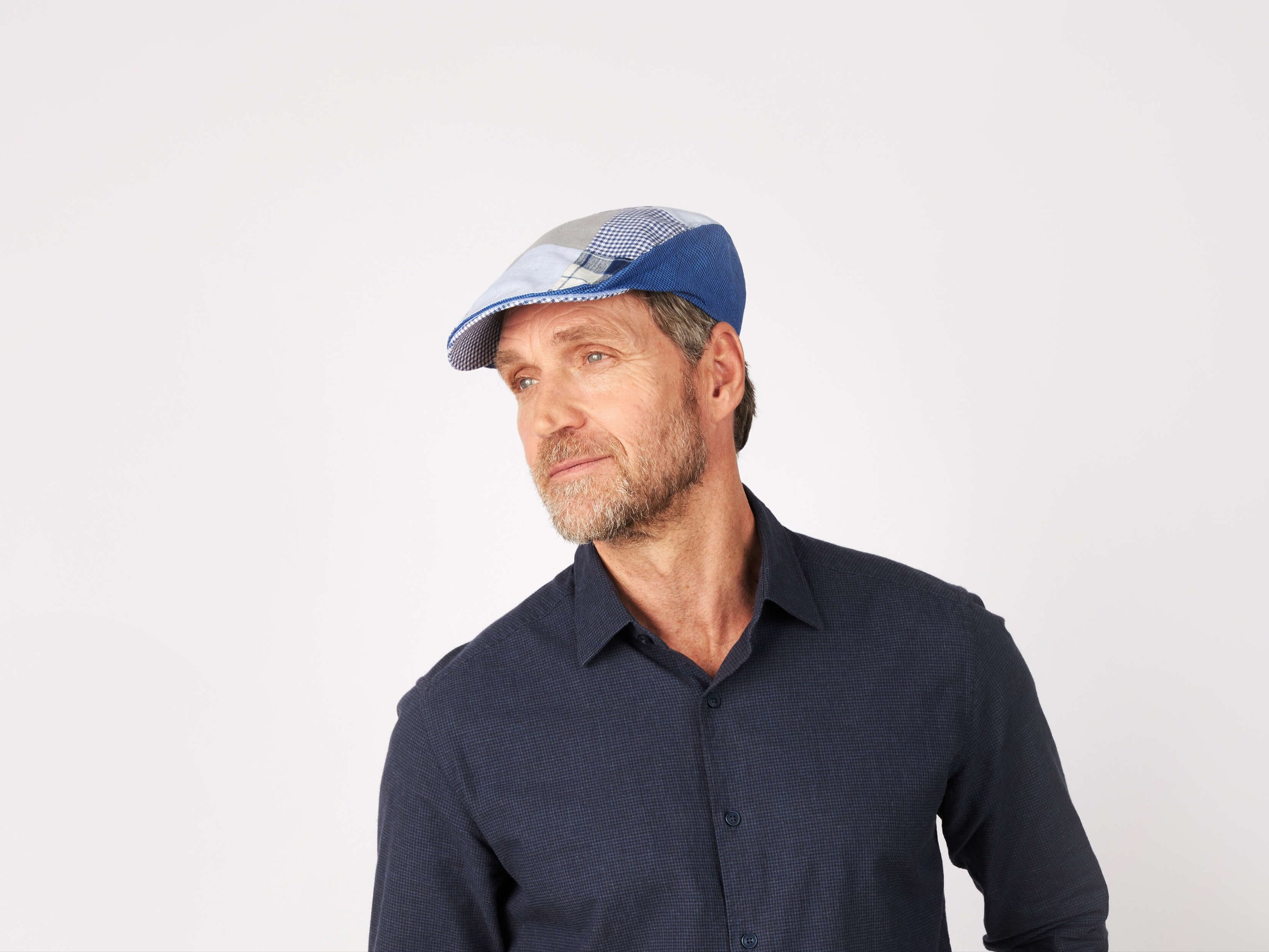 A middle-aged man with a beard wears a Hanna Hats Donegal Touring Cap Patchwork Grey/Blue Linen and a dark blue button-up shirt, looking thoughtfully to the side against a plain white background.