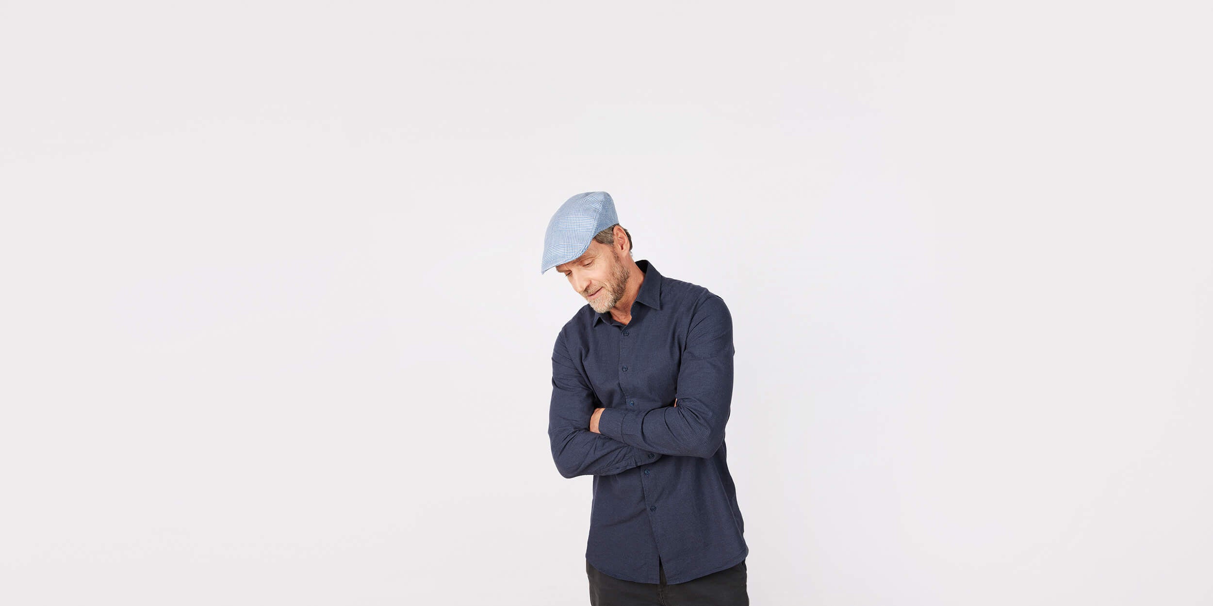 A man wearing a light blue flat cap and a dark blue button-up shirt stands with arms crossed, looking down, against a plain white background.