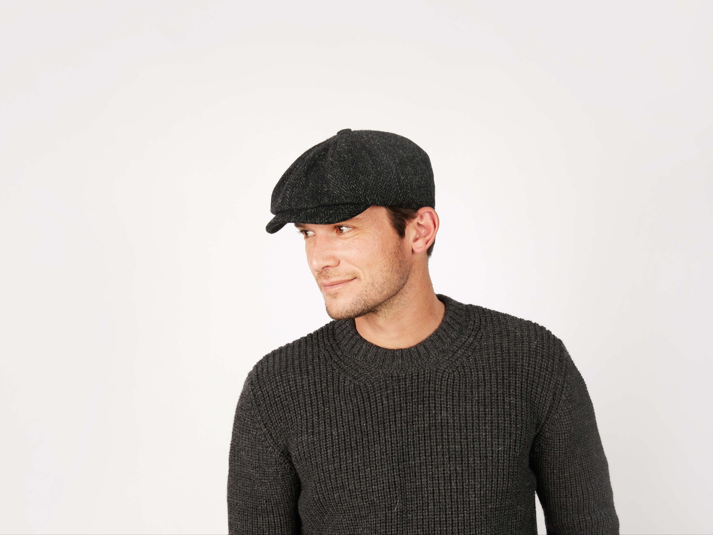 A man in a tailored fit dark sweater and a Hanna Hats JP Cap Tweed looks to his left against a plain white background.