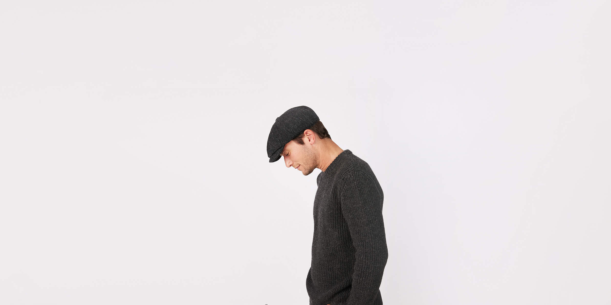 A man wearing a dark sweater and a flat cap is standing in profile against a plain white background, looking down with his hands in his pockets.