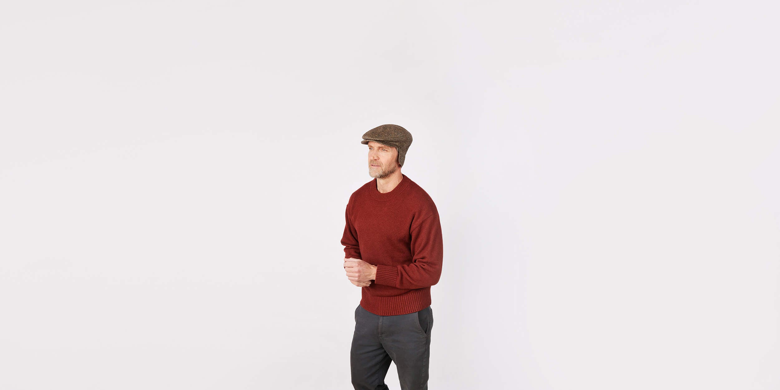 A man wearing a brown flat cap, a red sweater, and dark pants stands against a plain white background with his hands clasped in front of him.