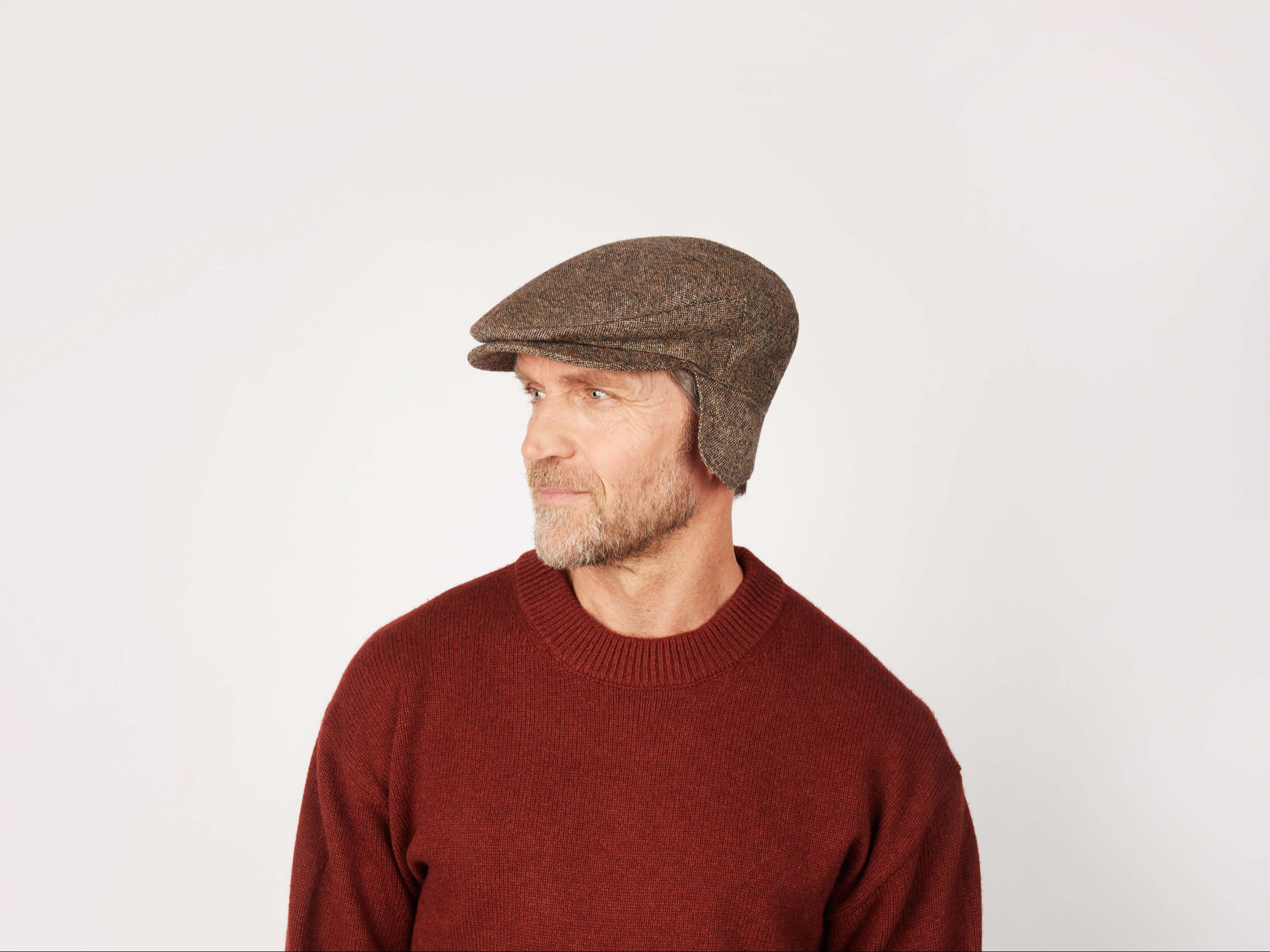 A bearded man wearing a Hanna Hats Vintage Cap Ear Flap Tweed and a maroon sweater looks to his left against a plain, light background.