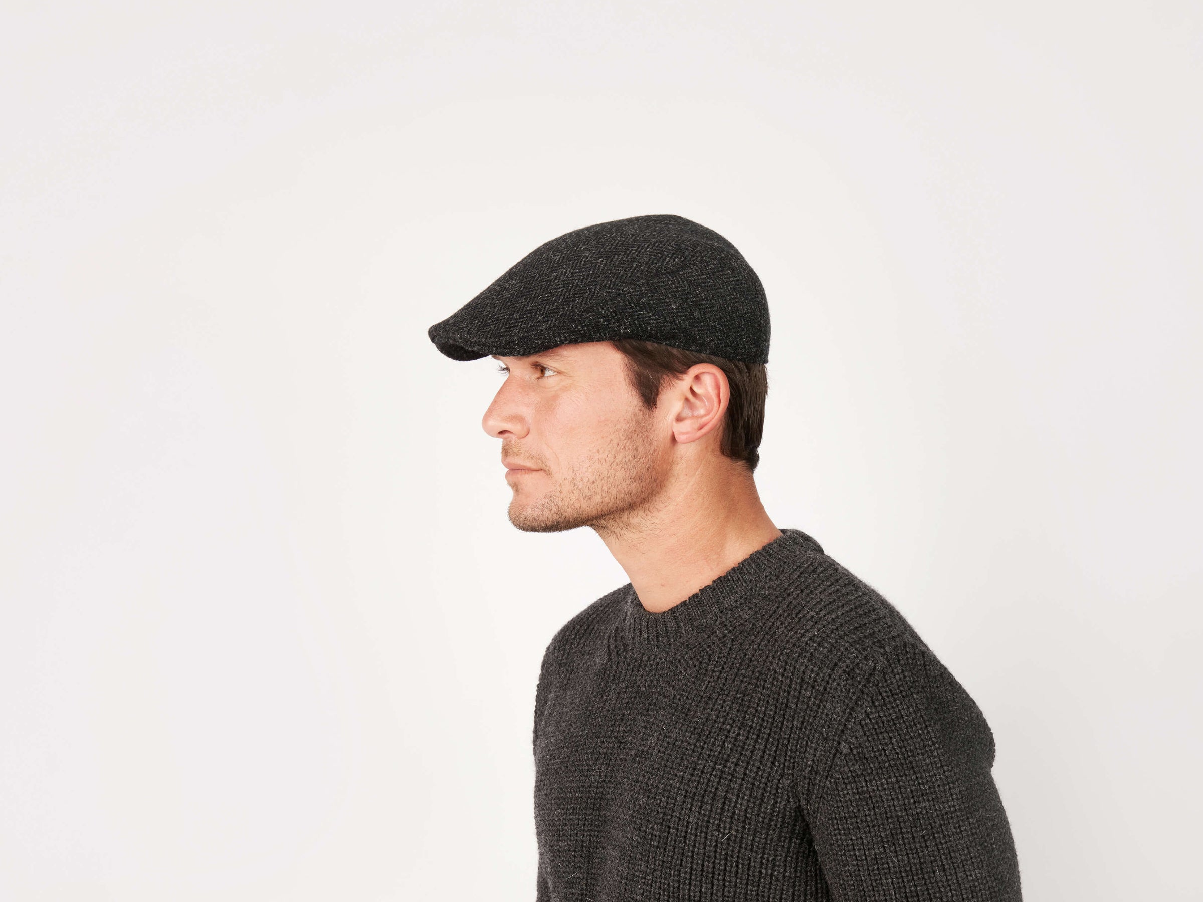 A man in a dark knit sweater and a tailored-fit Donegal Touring Cap Tweed by Hanna Hats is shown in profile against a plain white background.