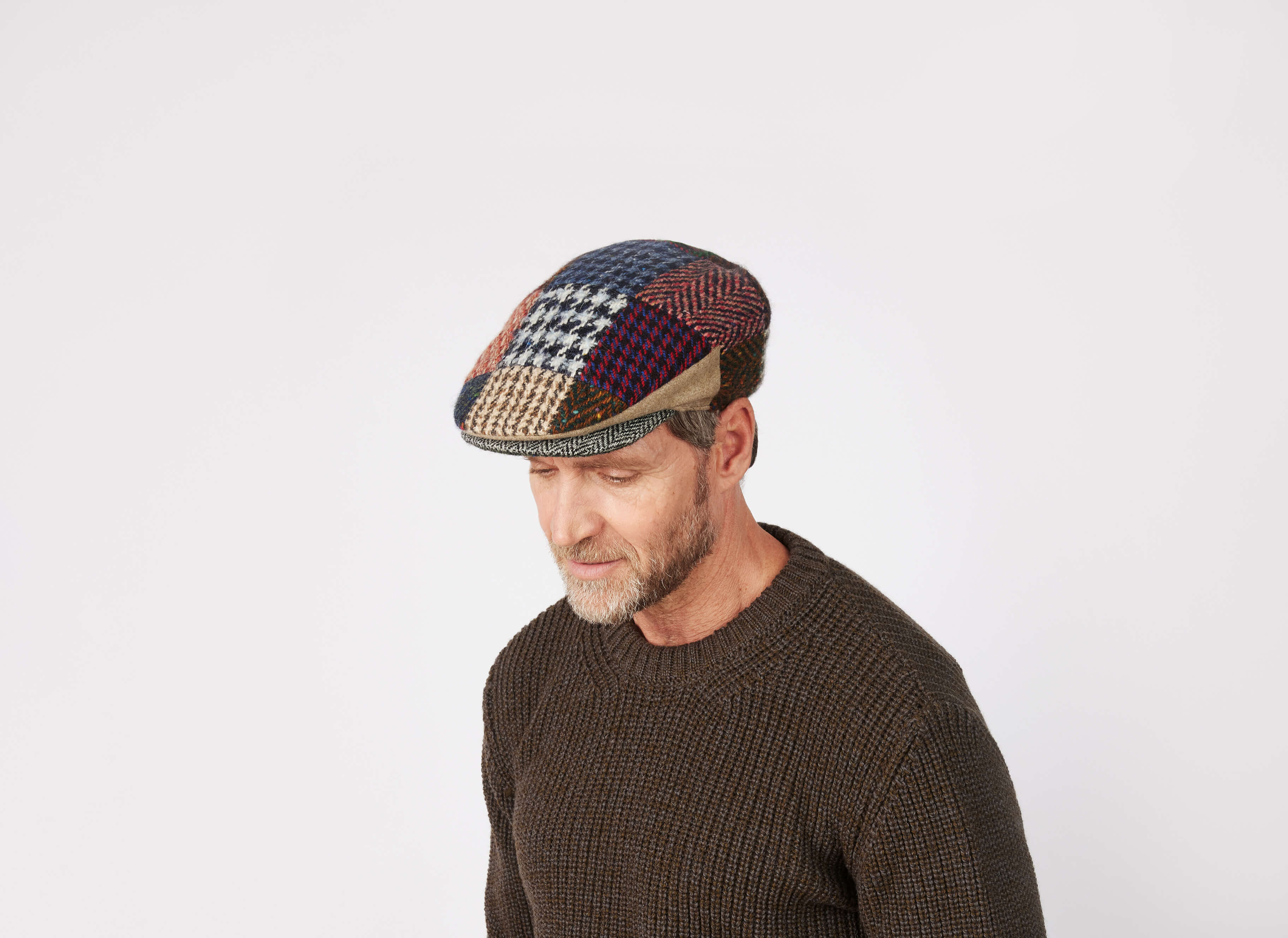 A man with a short beard wears a dark brown sweater and the Hanna Hats Vintage Cap Heavy Patchwork Tweed - LIMITED EDITION, looking down against a plain light background.