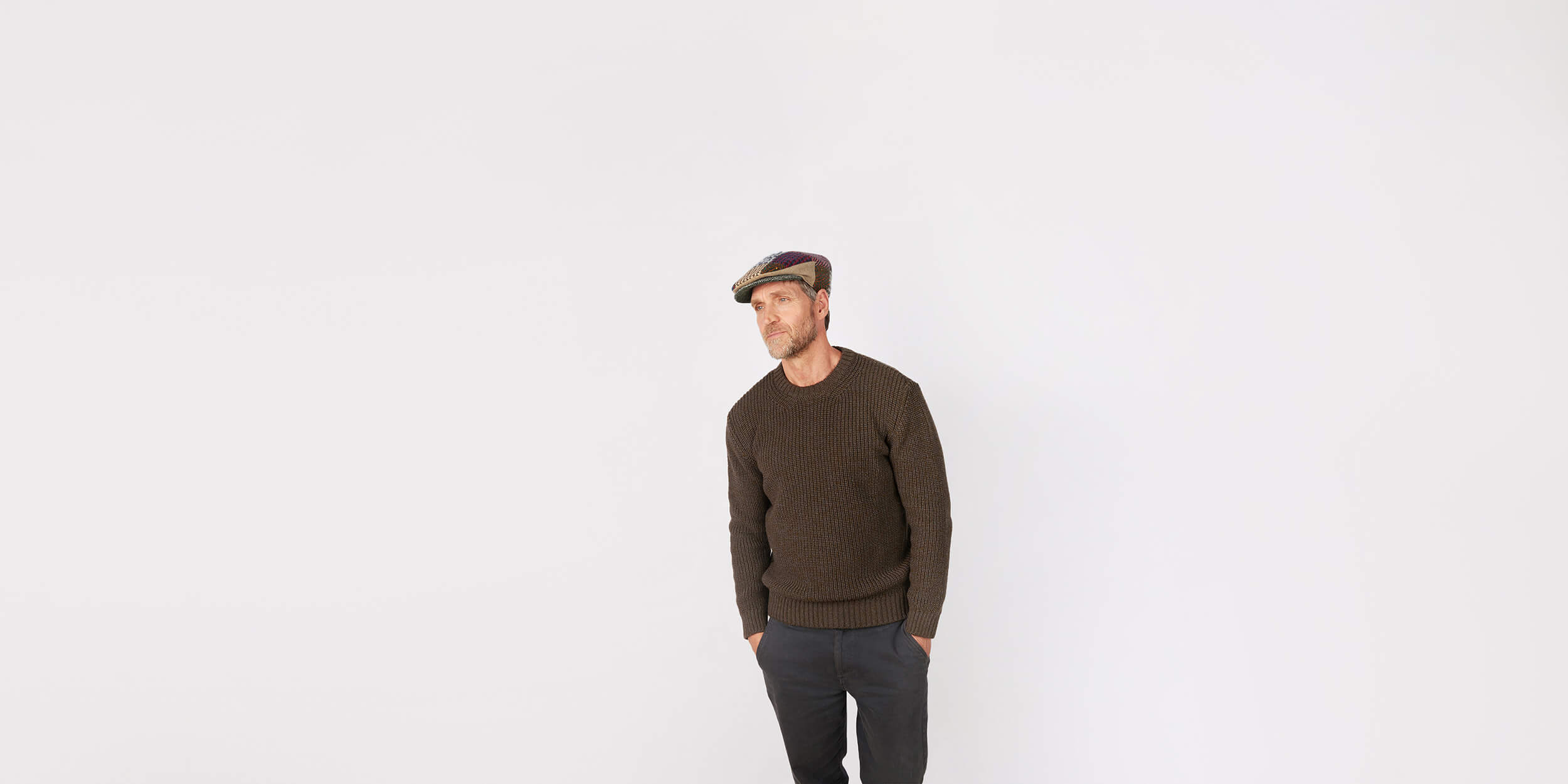 A man wearing a brown sweater, dark pants, and a flat cap stands against a plain white background with his hands in his pockets, looking slightly to the side.