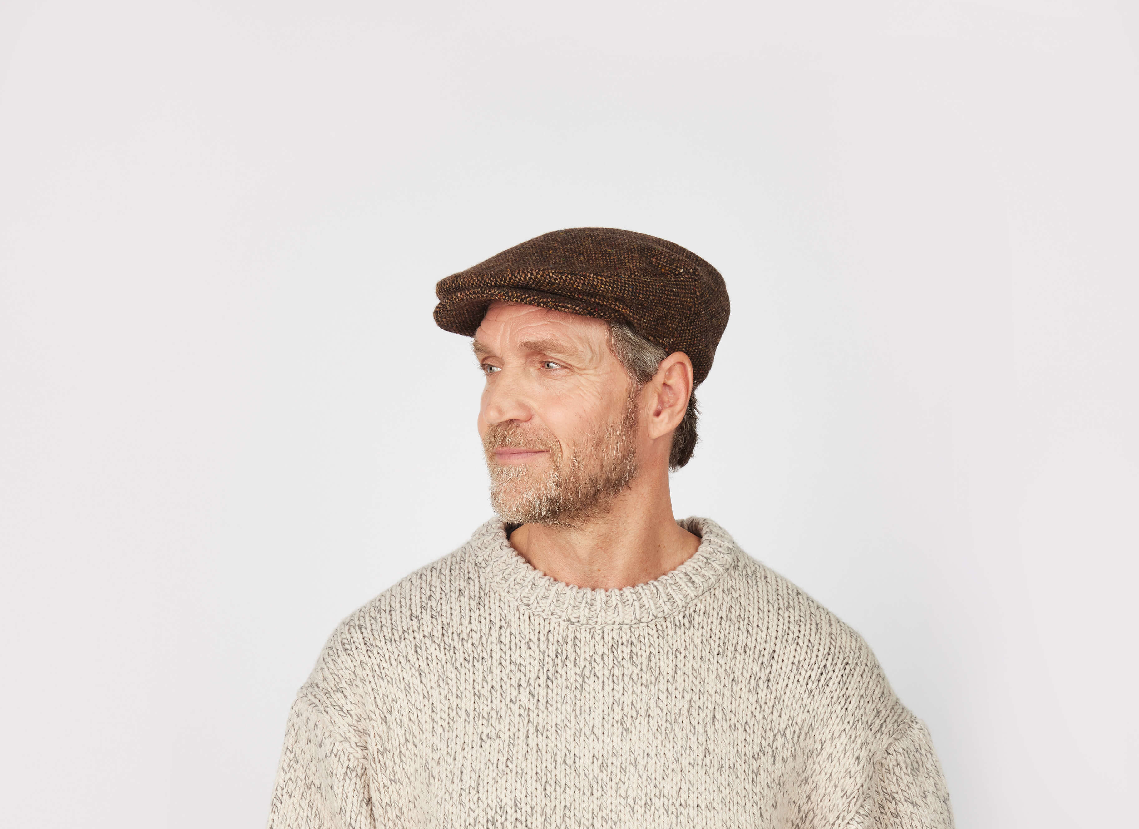 A middle-aged man with a beard wears the Hanna Hats Vintage Cap Tweed and a chunky knit sweater, looking to his left against a plain light background.