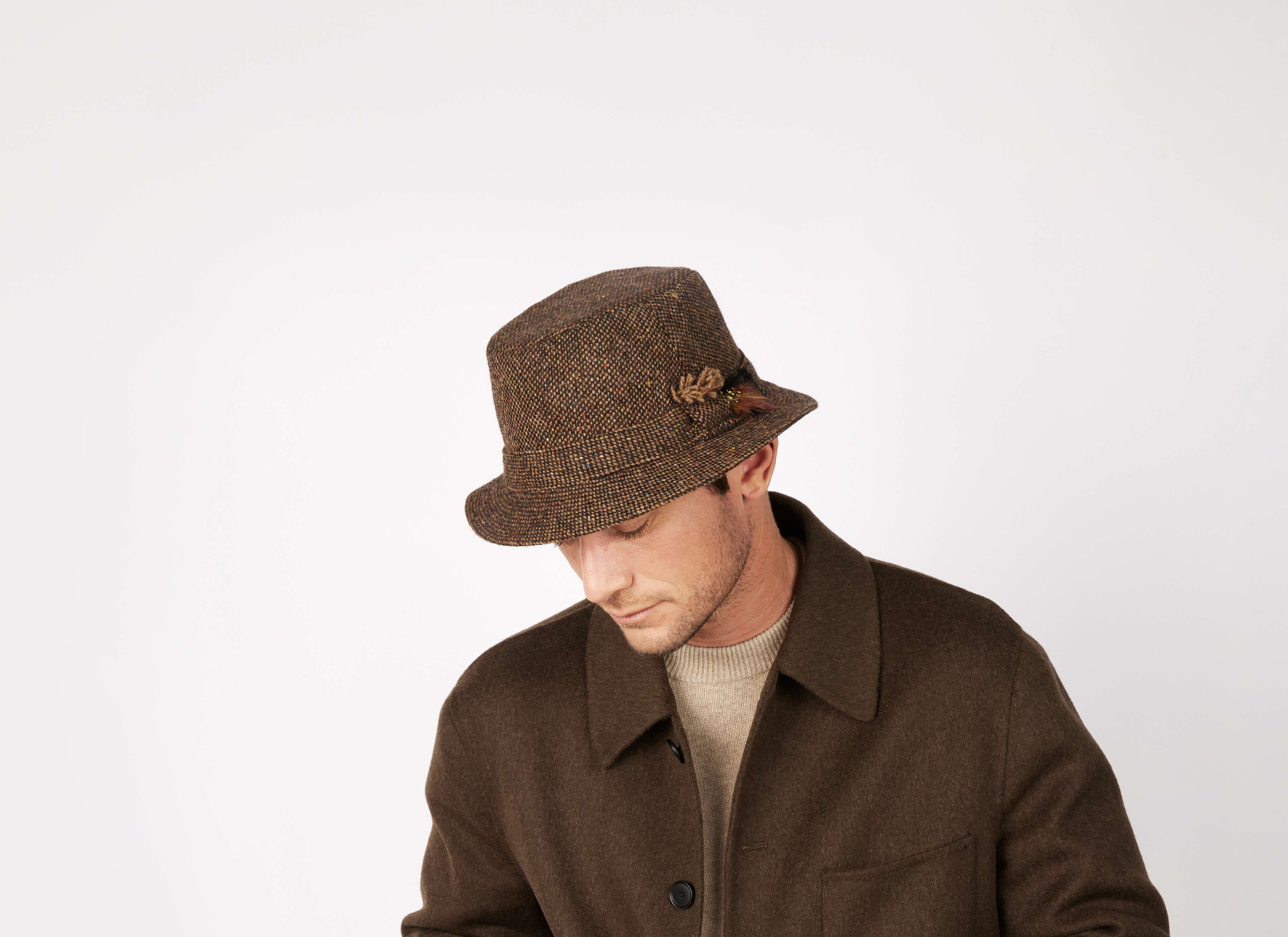 A man in a Hanna Hats Walking Hat Tweed in brown with decorative pin and dark brown coat over a beige sweater looks down against a plain white background.