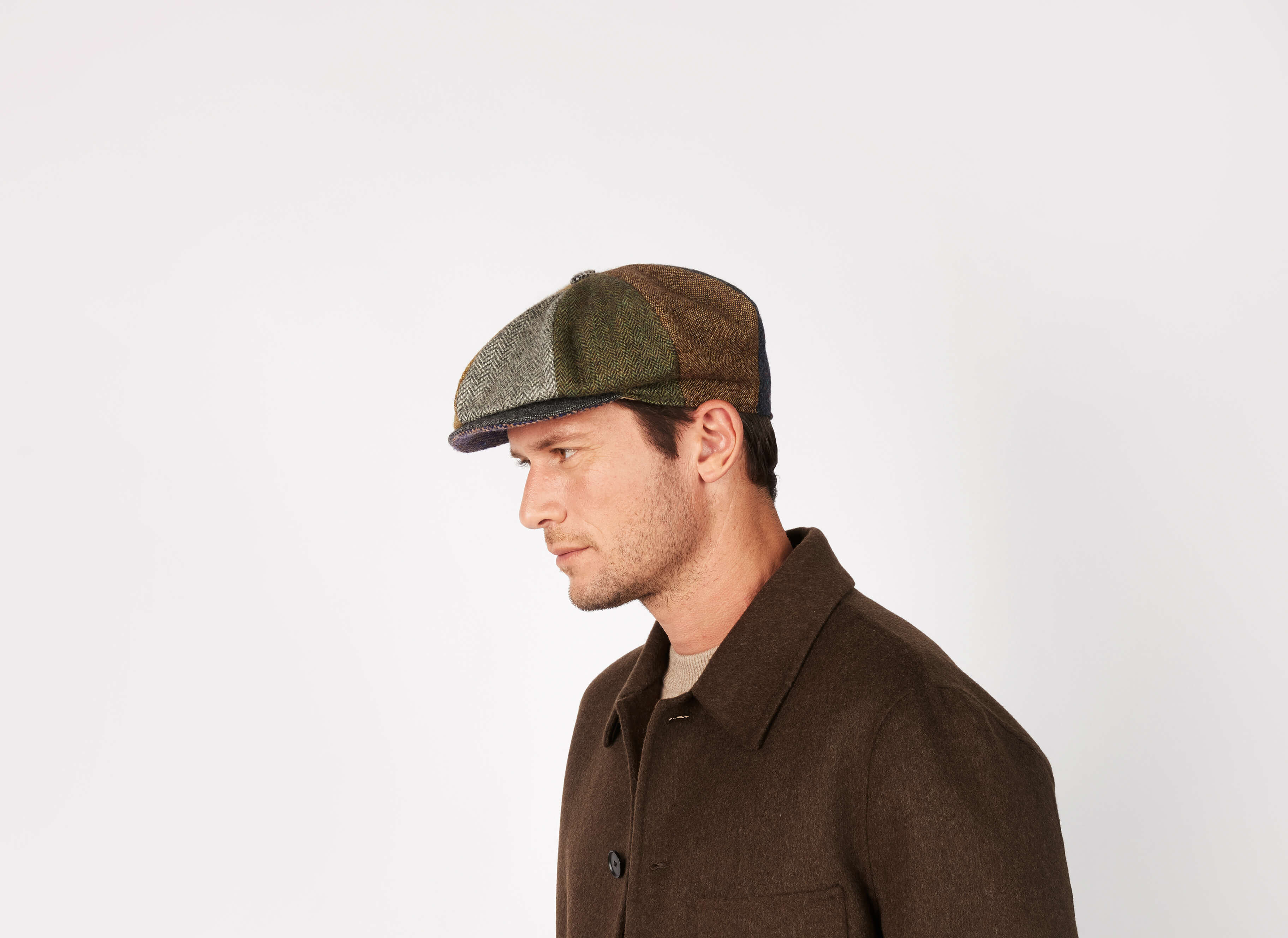 A man wearing a brown wool coat and a Hanna Hats Eight Piece Cap Patchwork Tweed, handcrafted in Ireland, looks to the left against a plain white background.