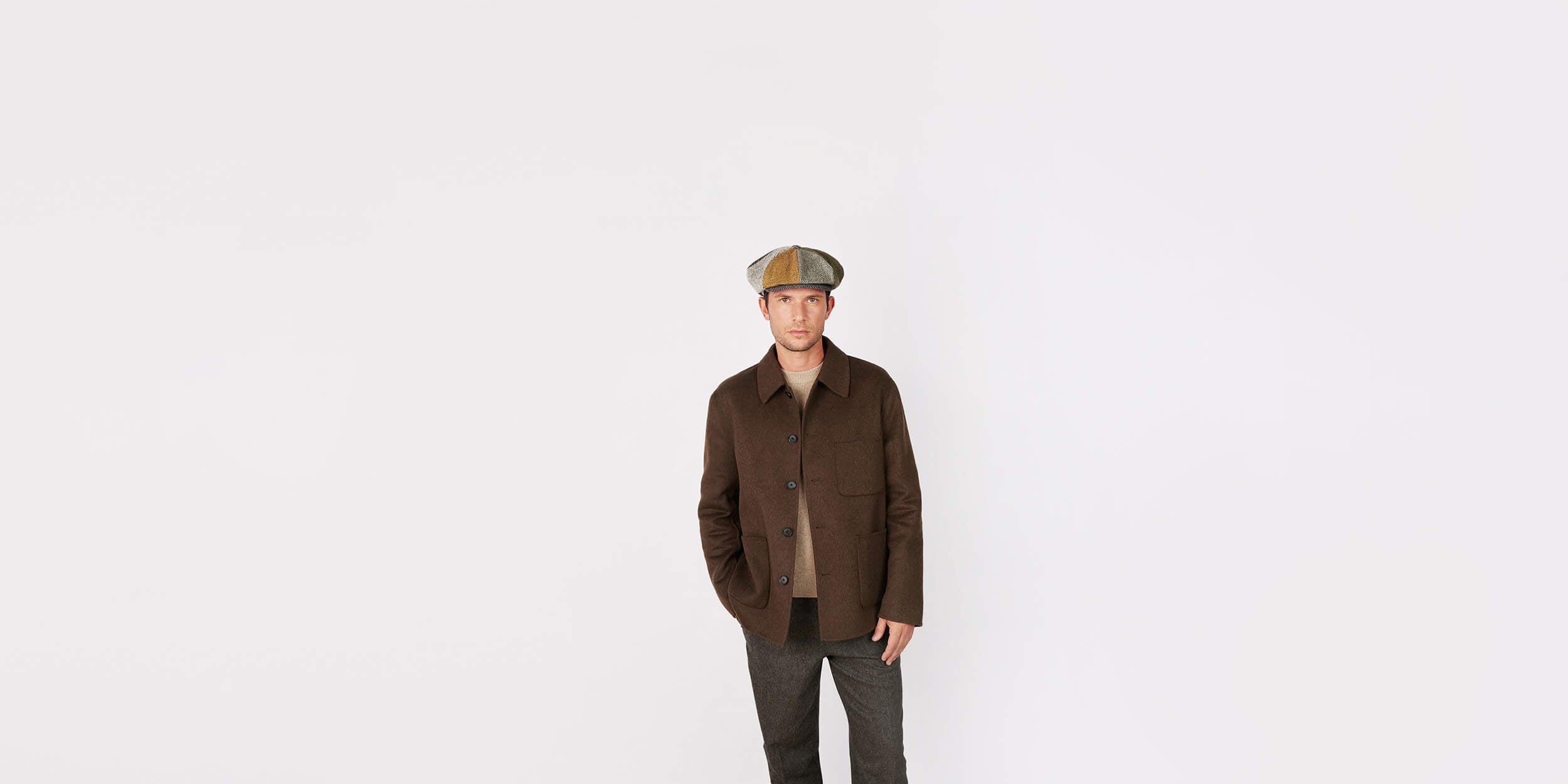 A man wearing a brown buttoned jacket, gray pants, and a flat cap stands against a plain white background, facing forward with his hands in his jacket pockets.