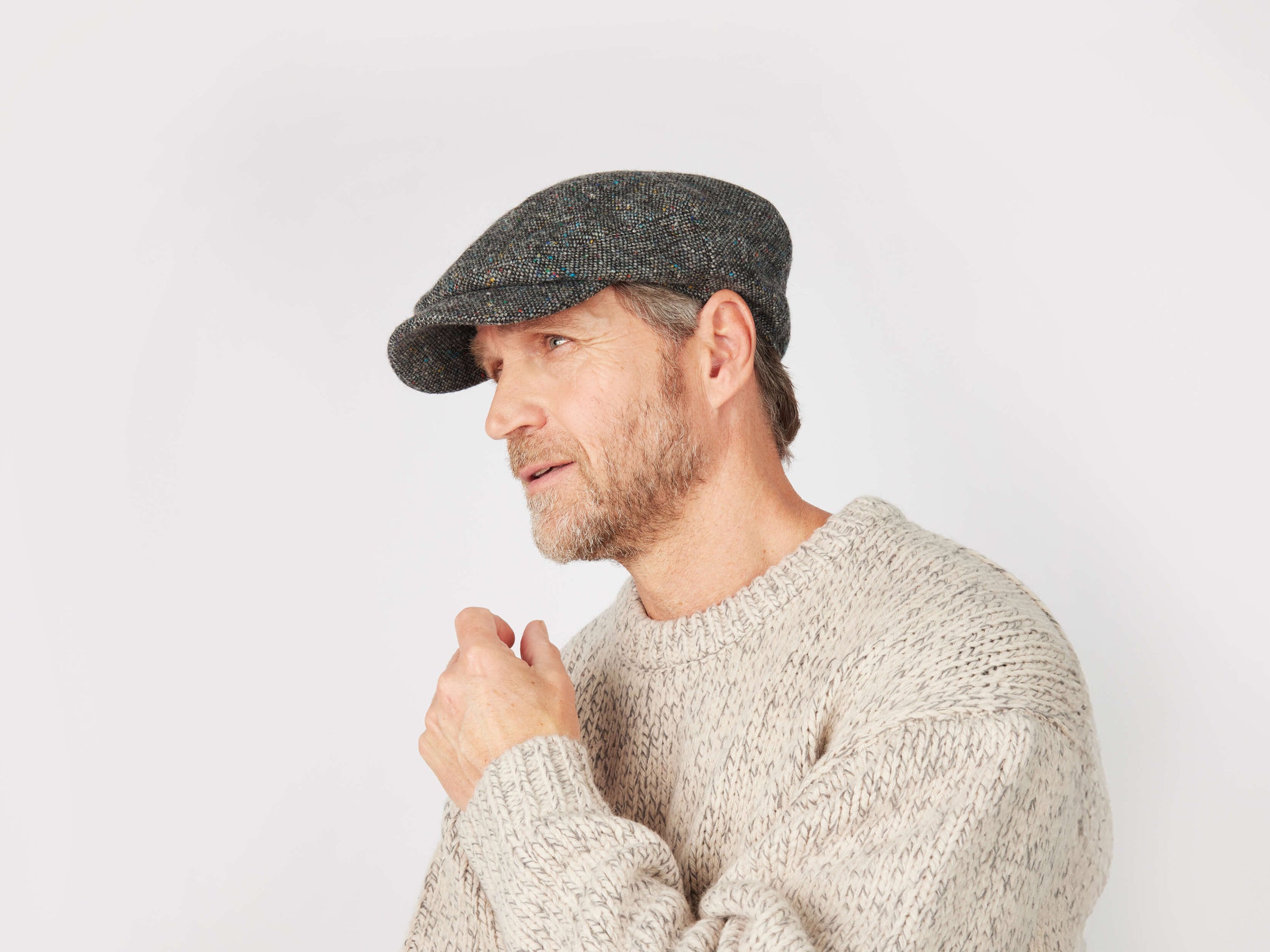 A middle-aged man in a Daithi Cap Tweed by Hanna Hats and a beige knit sweater looks thoughtful against a plain white background.