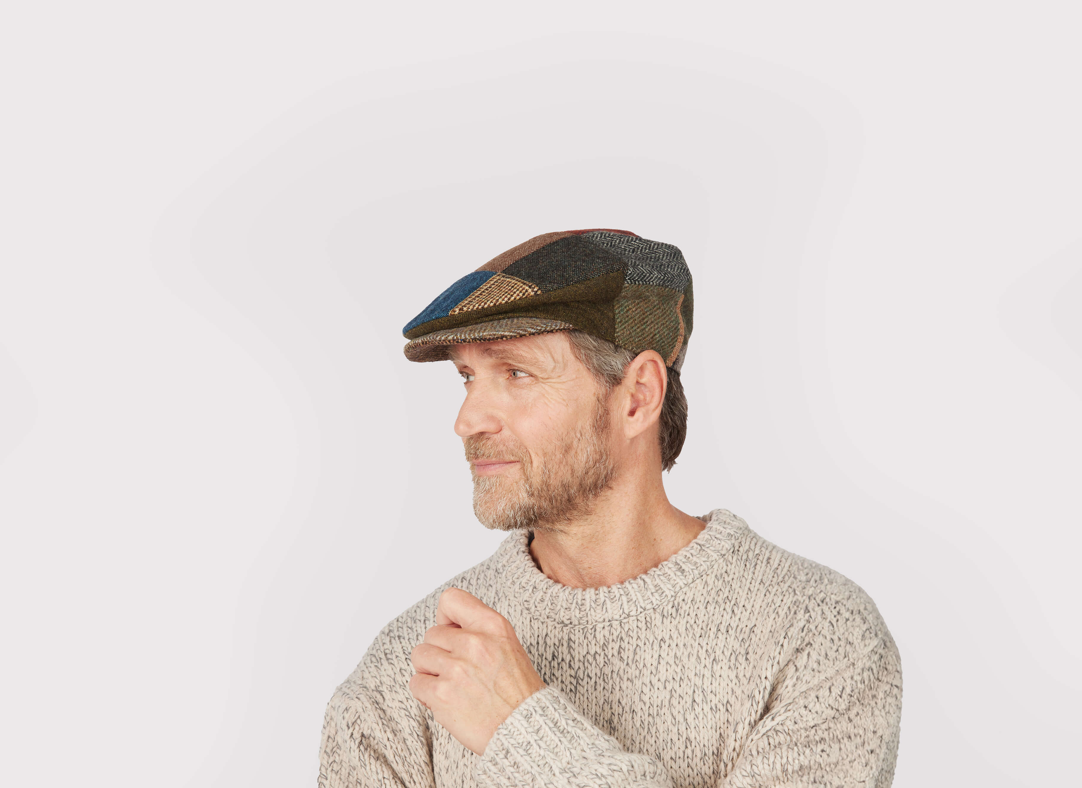 A middle-aged bearded man wears the Hanna Hats Vintage Cap Patchwork Tweed and a light knit sweater, glancing left against a plain background. The vintage cap brings classic charm to his casual look.