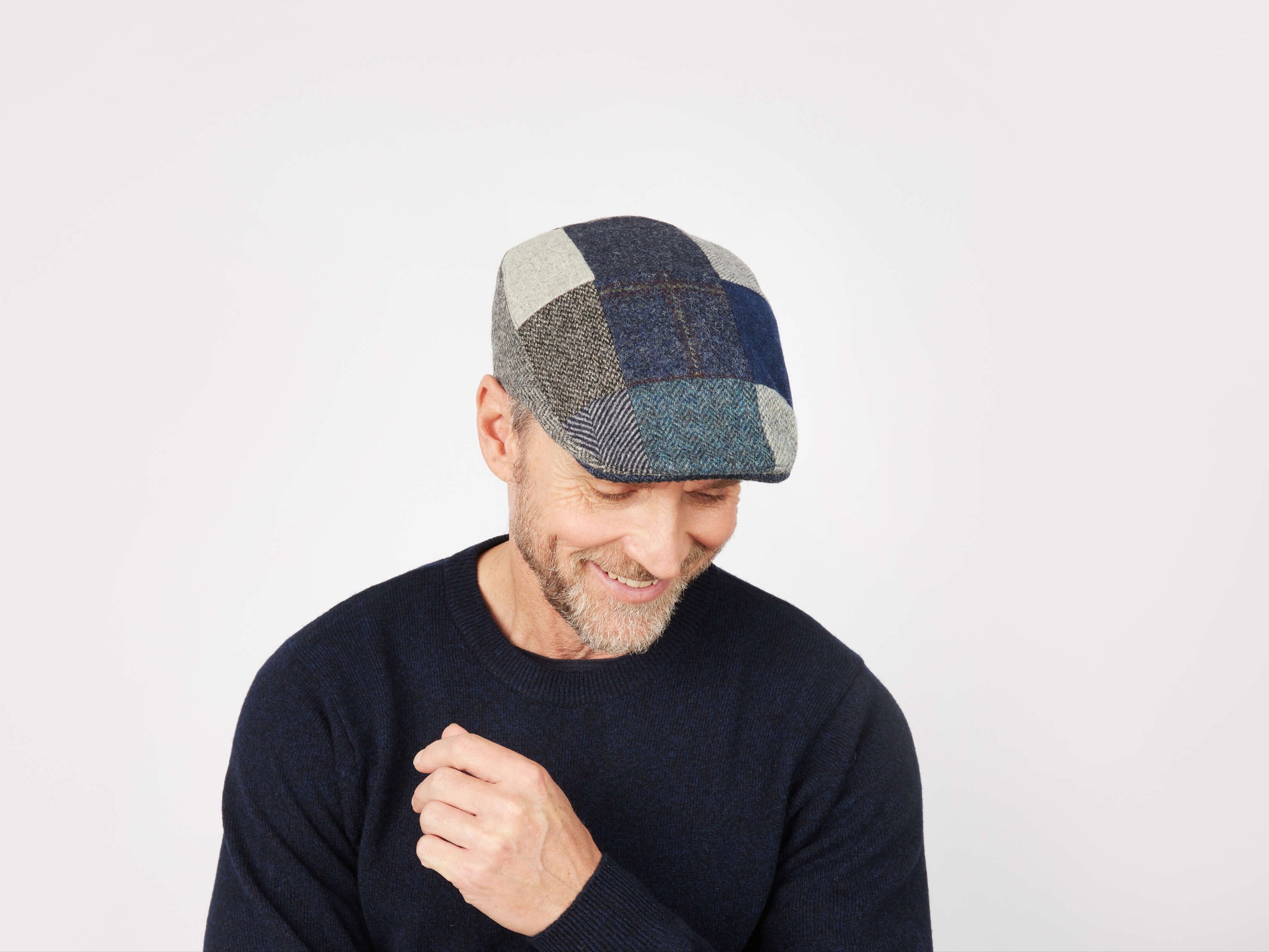 A man wearing a dark sweater and the Hanna Hats Donegal Touring Cap Patchwork Grey/Blue Tweed smiles while looking down against a plain white background.