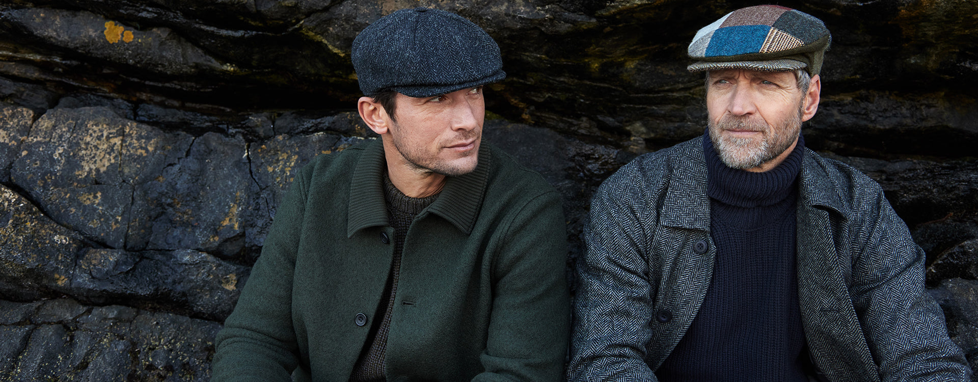 Two men wearing flat caps and jackets sit in front of a rocky background, looking thoughtfully into the distance. One man has a gray beard and patchwork cap; the other has a dark cap and is clean-shaven.