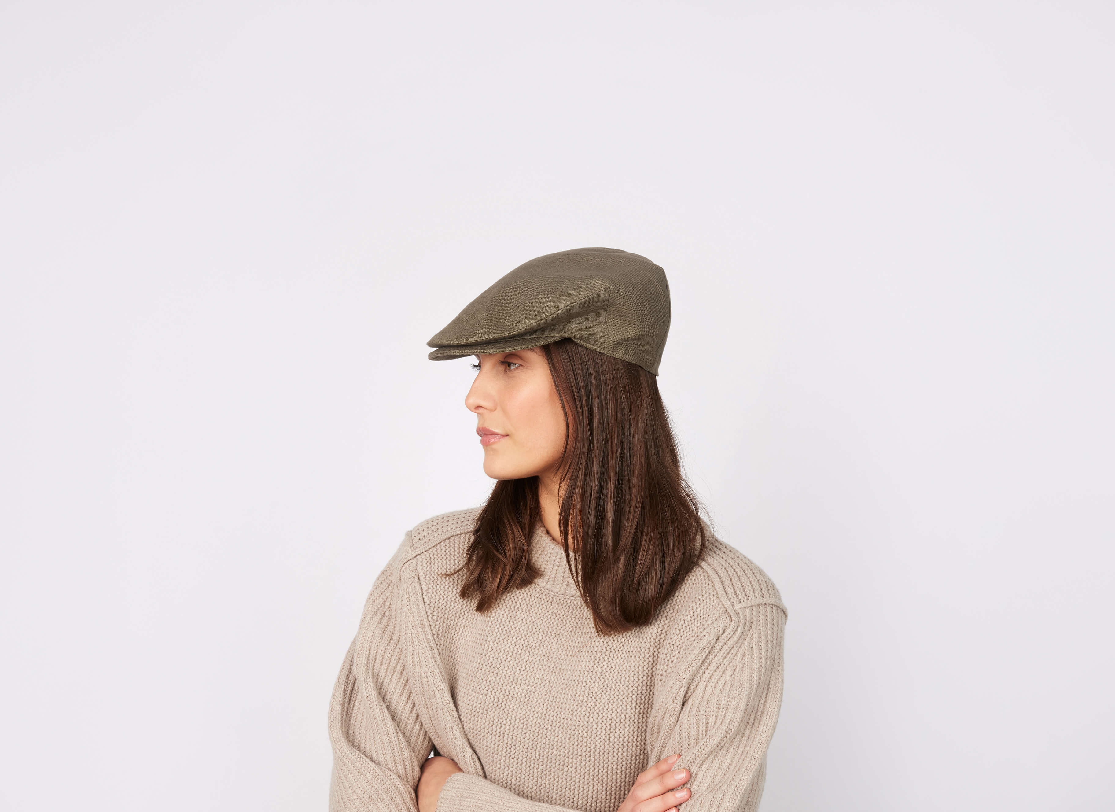 A woman with straight brown hair wears a beige sweater and the Hanna Hats Vintage Cap Linen, standing against a white background and looking to her left with arms crossed.