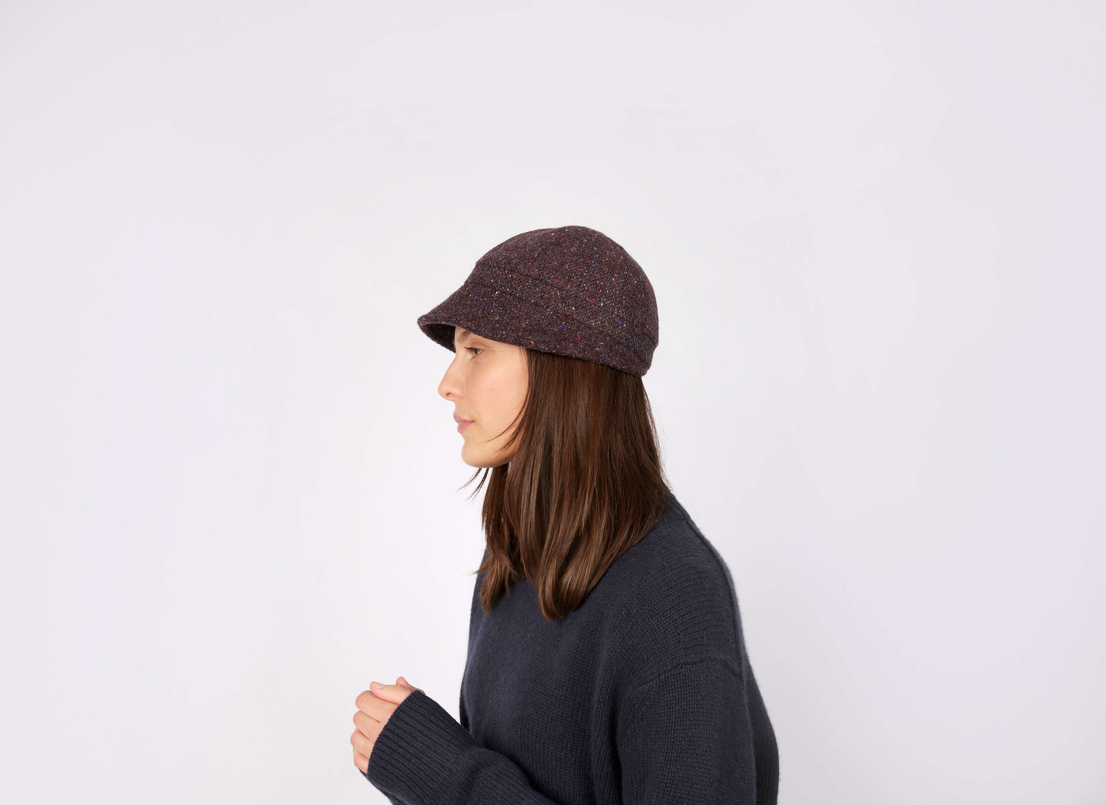 A woman with straight brown hair in a dark knit sweater and a Hanna Hats Slieve League Hat Tweed stands in profile against a plain white background.