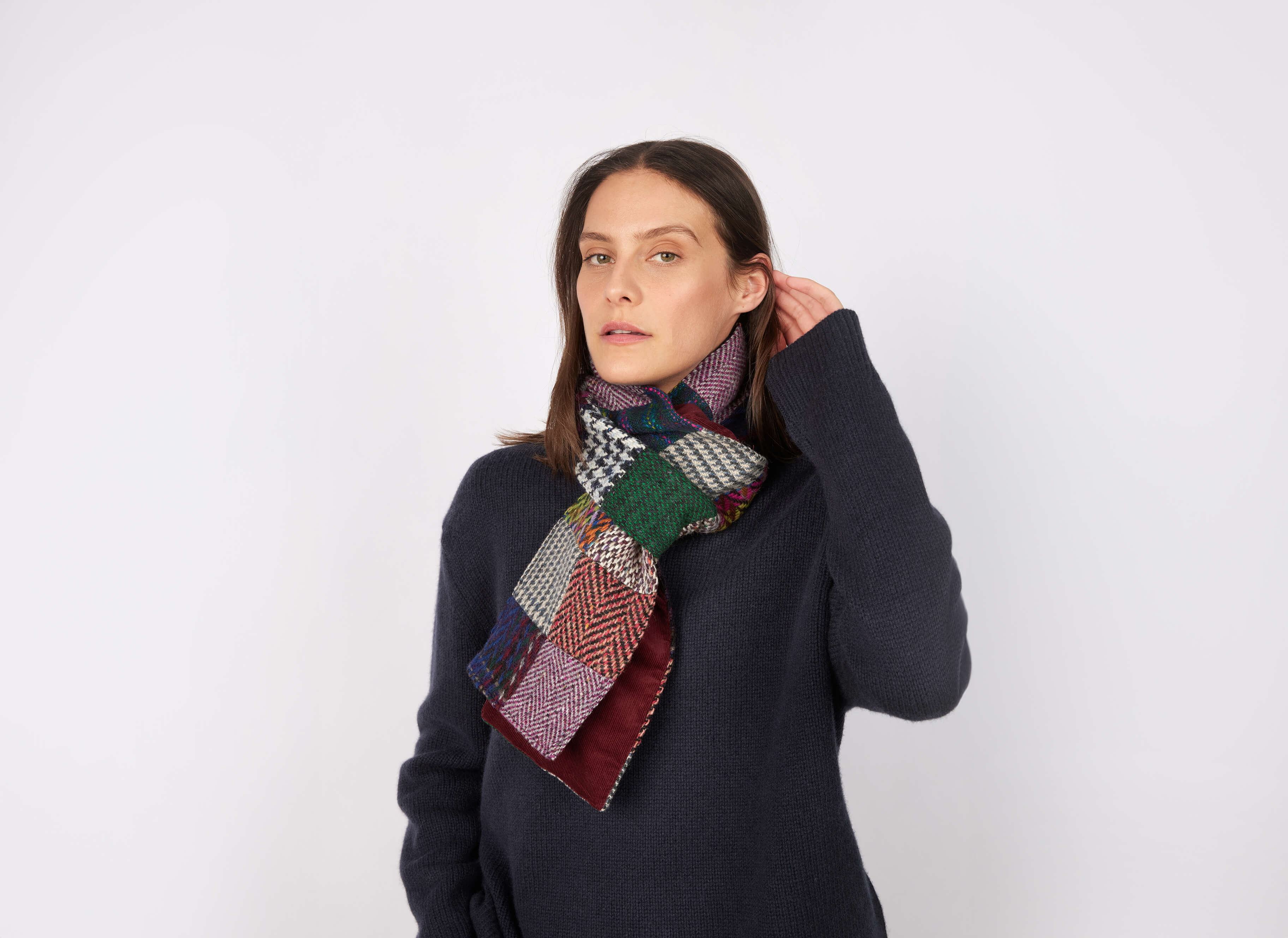 A woman with straight brown hair wears a dark sweater and the Hanna Hats Scarf Heavy Patchwork Tweed / Corduroy - LIMITED EDITION, standing against a white background, looking at the camera with a neutral expression, one hand touching her head.