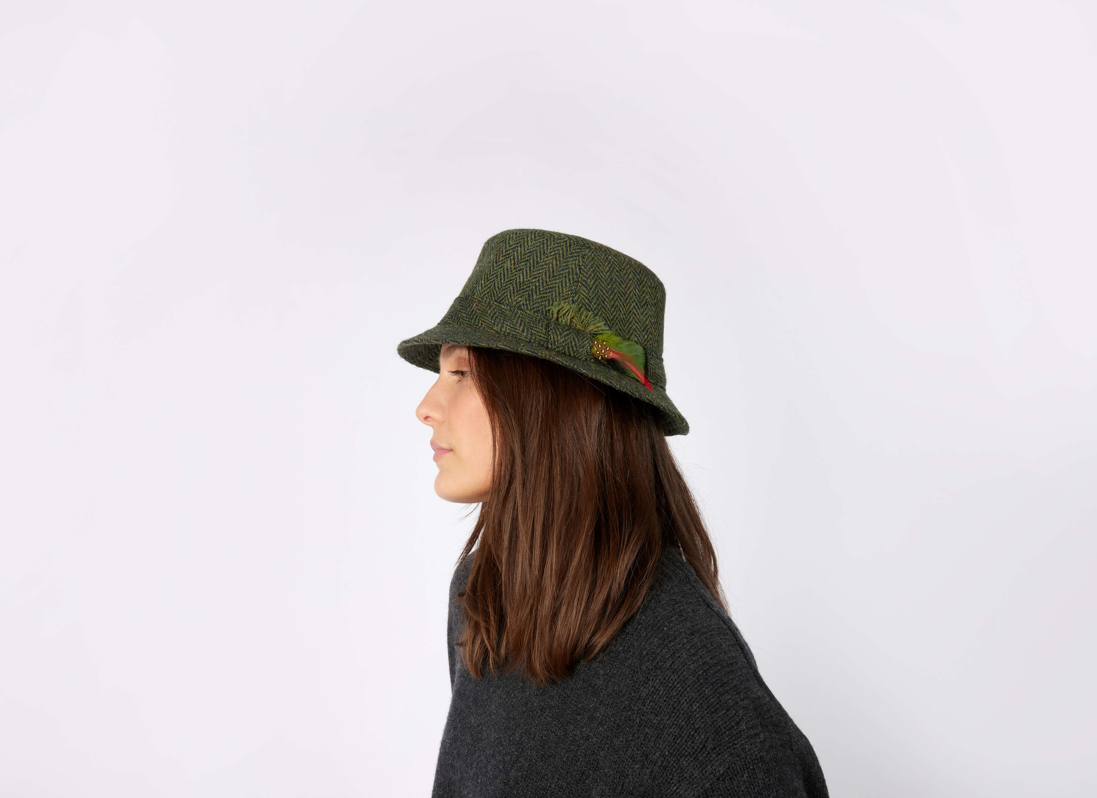 A person with straight brown hair, seen in profile against a white background, wears the Hanna Hats Walking Hat Tweed—a green Irish hat with a feather, crafted from authentic Irish tweed.