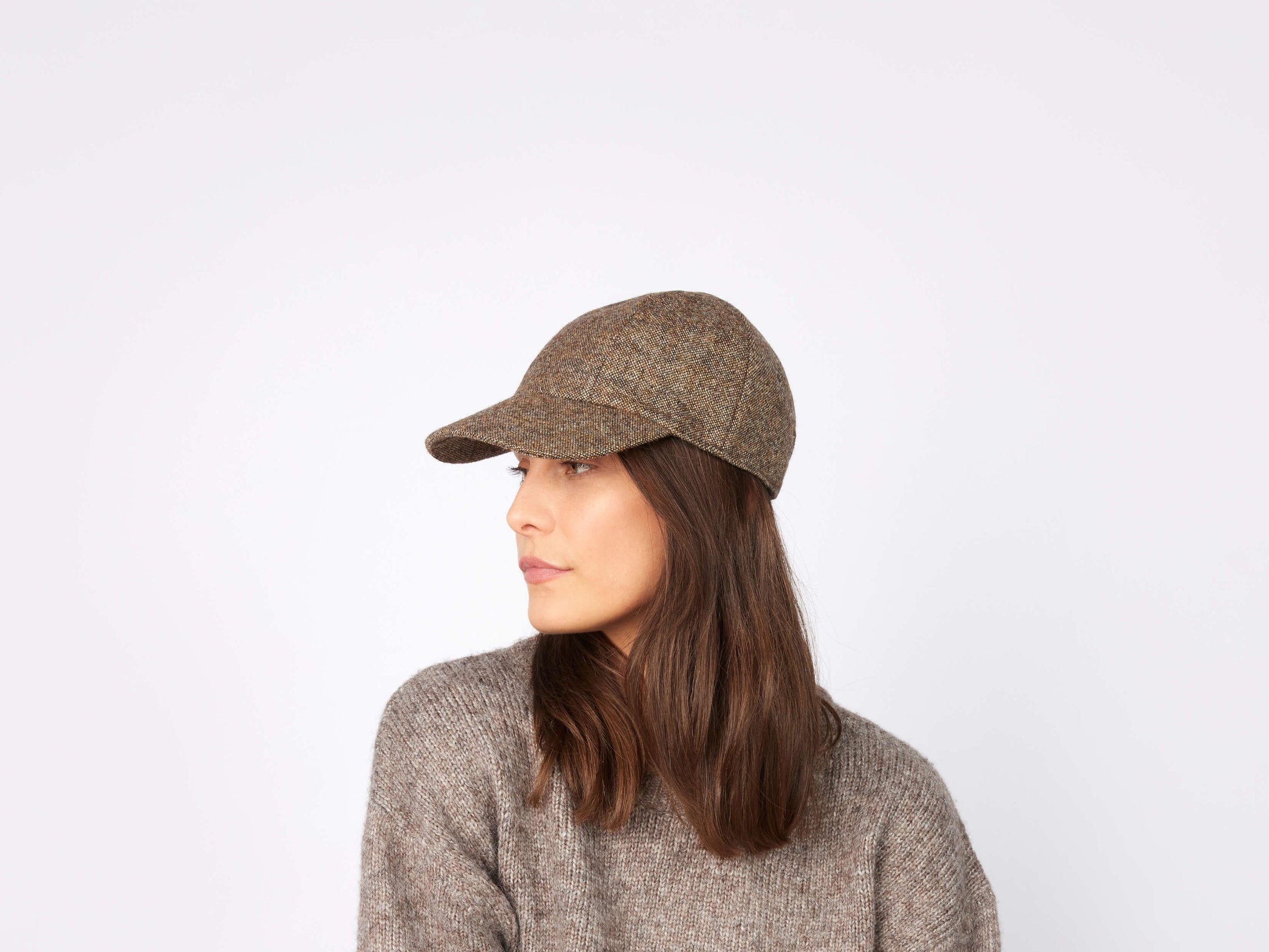 A woman with long brown hair wears a Hanna Hats Baseball Cap Tweed and a grey knit sweater, looking to her left against a plain light background.
