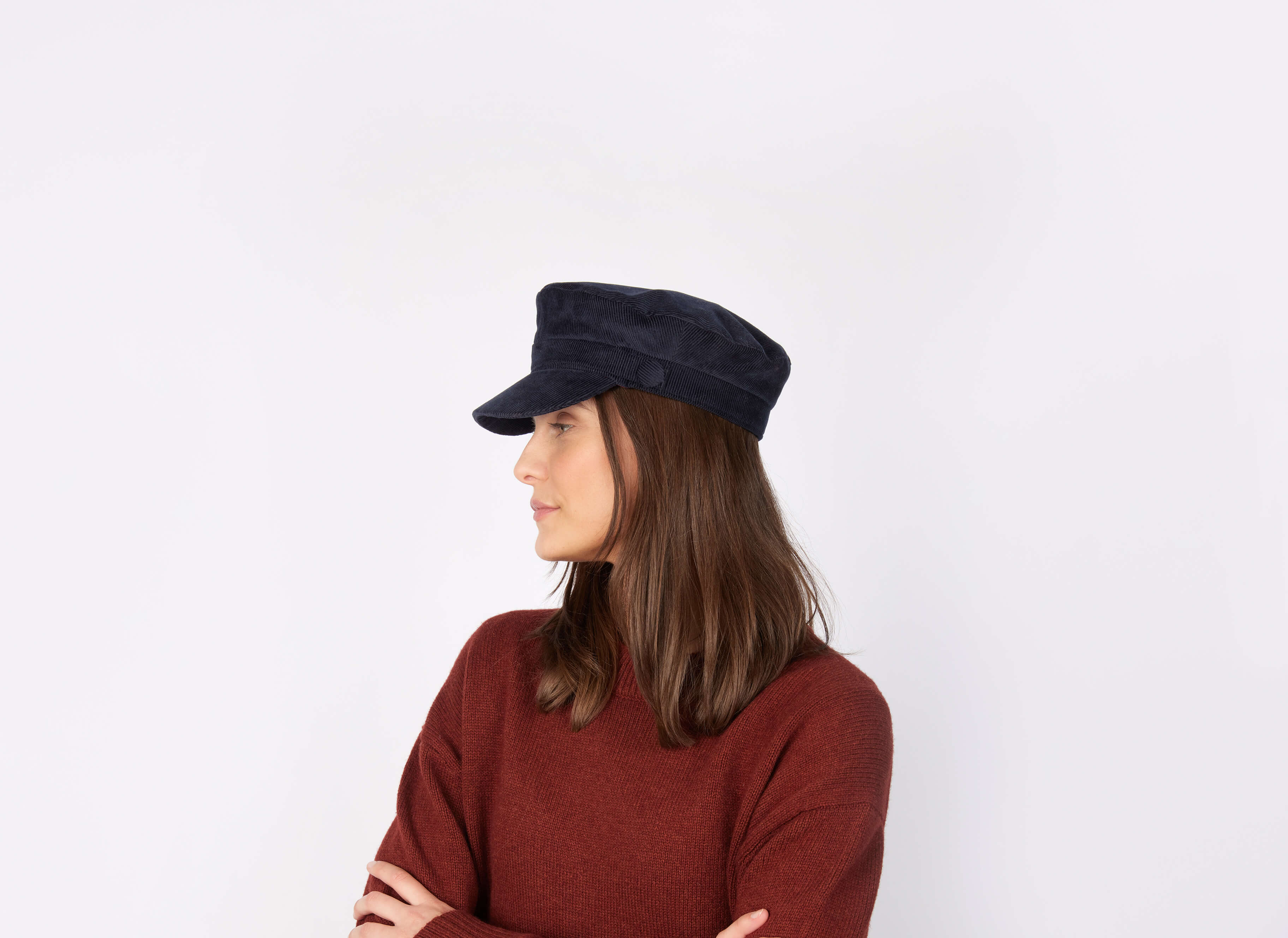A person with long brown hair wears a dark navy Hanna Hats Wild Atlantic Cap Cord with a cotton cord and a rust-colored sweater, standing arms crossed and facing left against a plain white background.