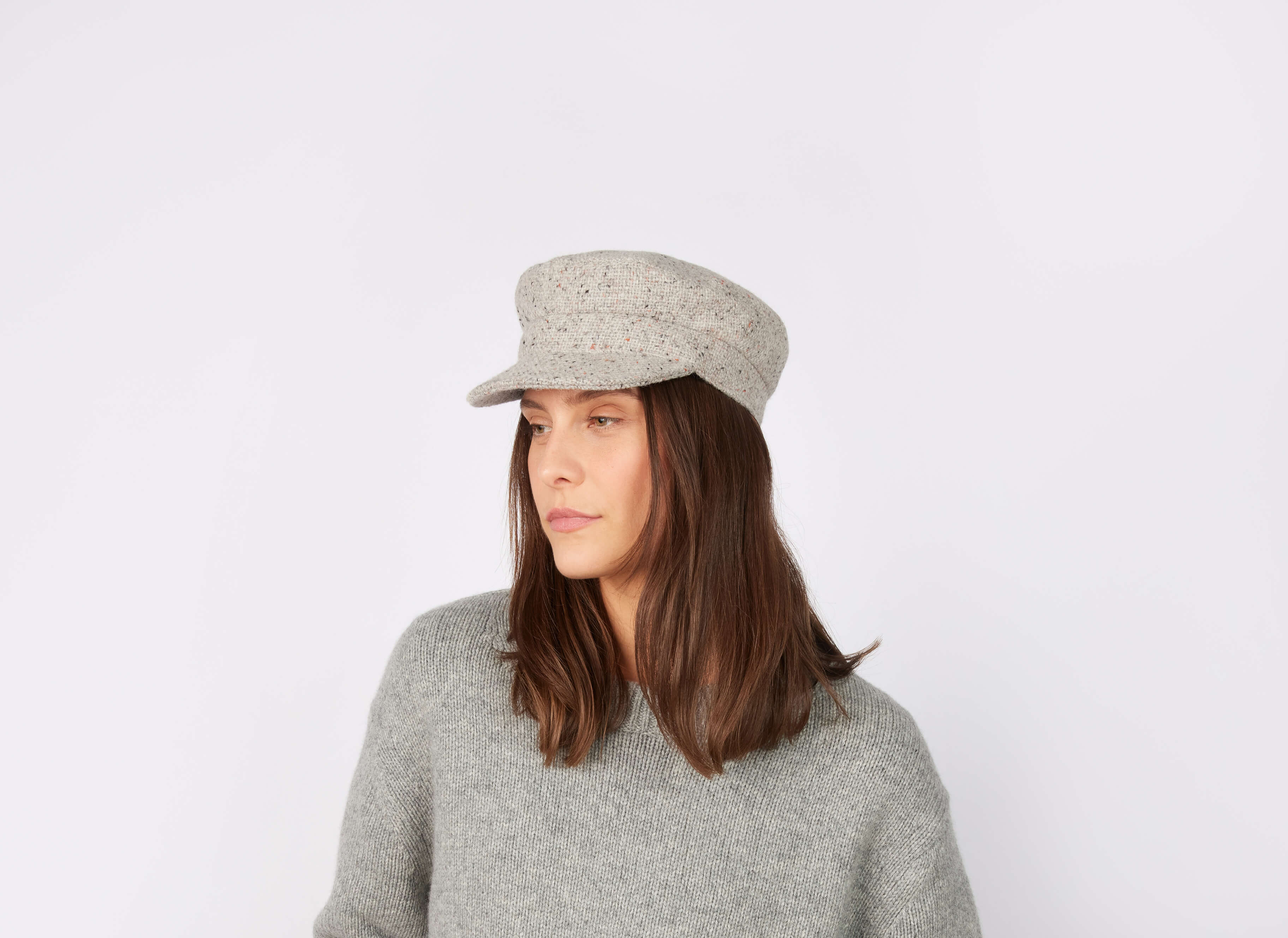A woman with straight brown hair wears a light gray Skipper Cap Tweed by Hanna Hats and a matching gray sweater, standing against a plain white background and gazing slightly to the side with a neutral expression.