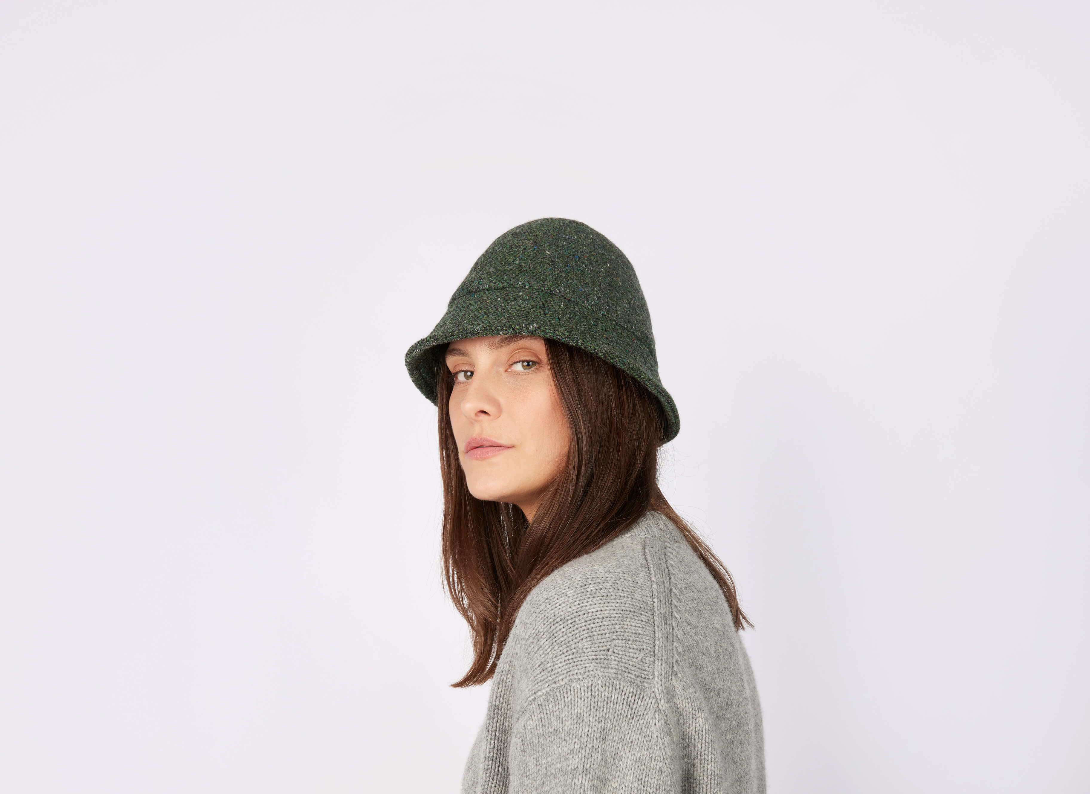 Woman with long brown hair wears a dark green Hanna Hats Eske Travel Hat Tweed and a gray tweed sweater, glancing over her shoulder against a plain white background.