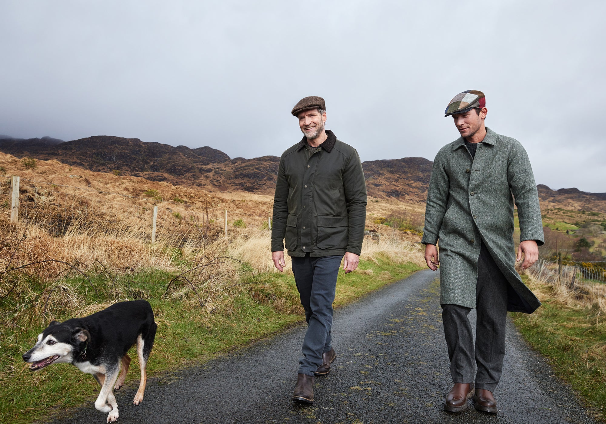 Two men wearing coats and flat caps walk along a rural road with a black and white dog. Rolling hills and cloudy skies form the scenic background.