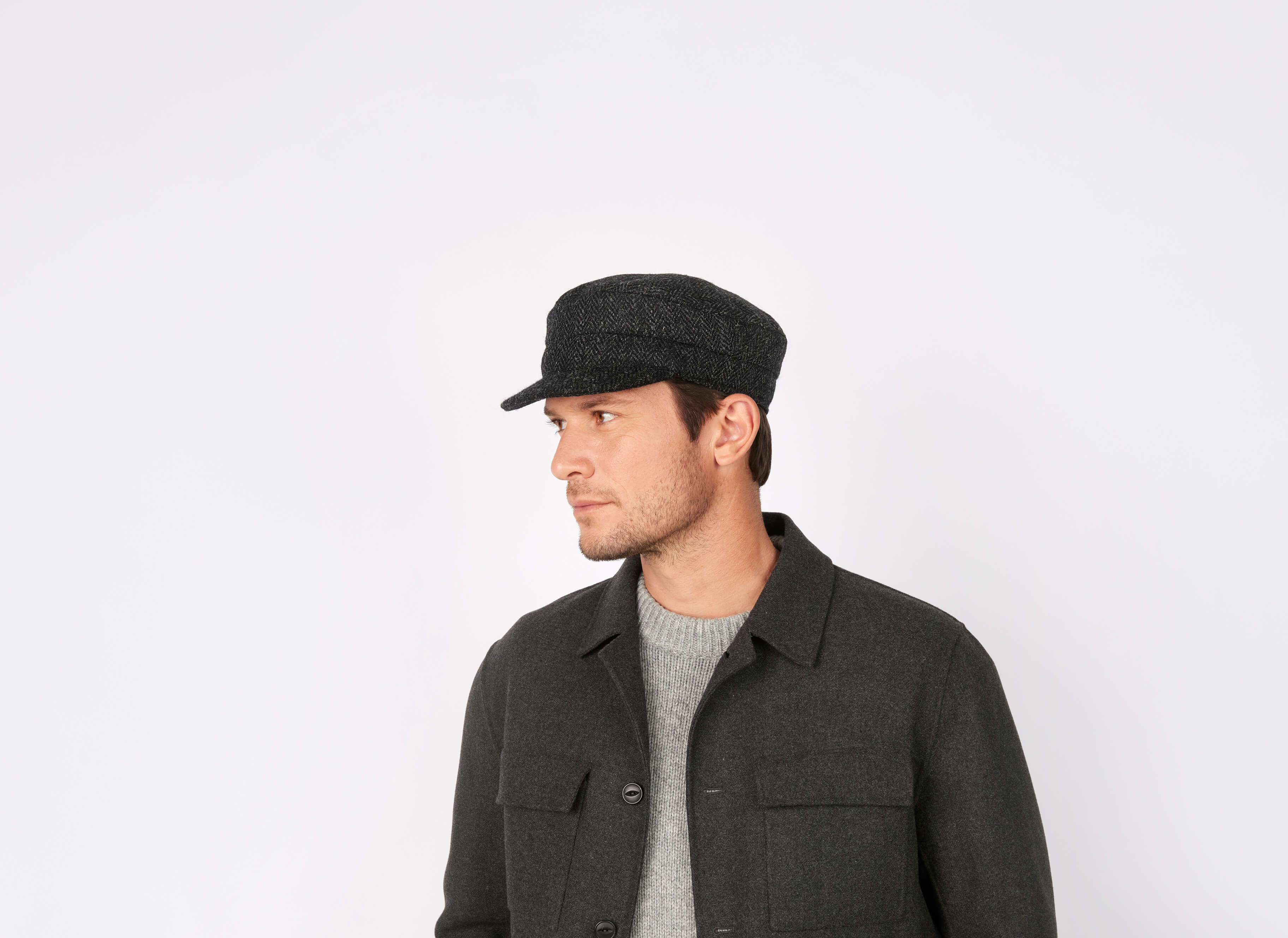A man wearing a Hanna Hats Skipper Cap Tweed in gray, a gray sweater, and a dark gray jacket looks to his left against a plain white background.