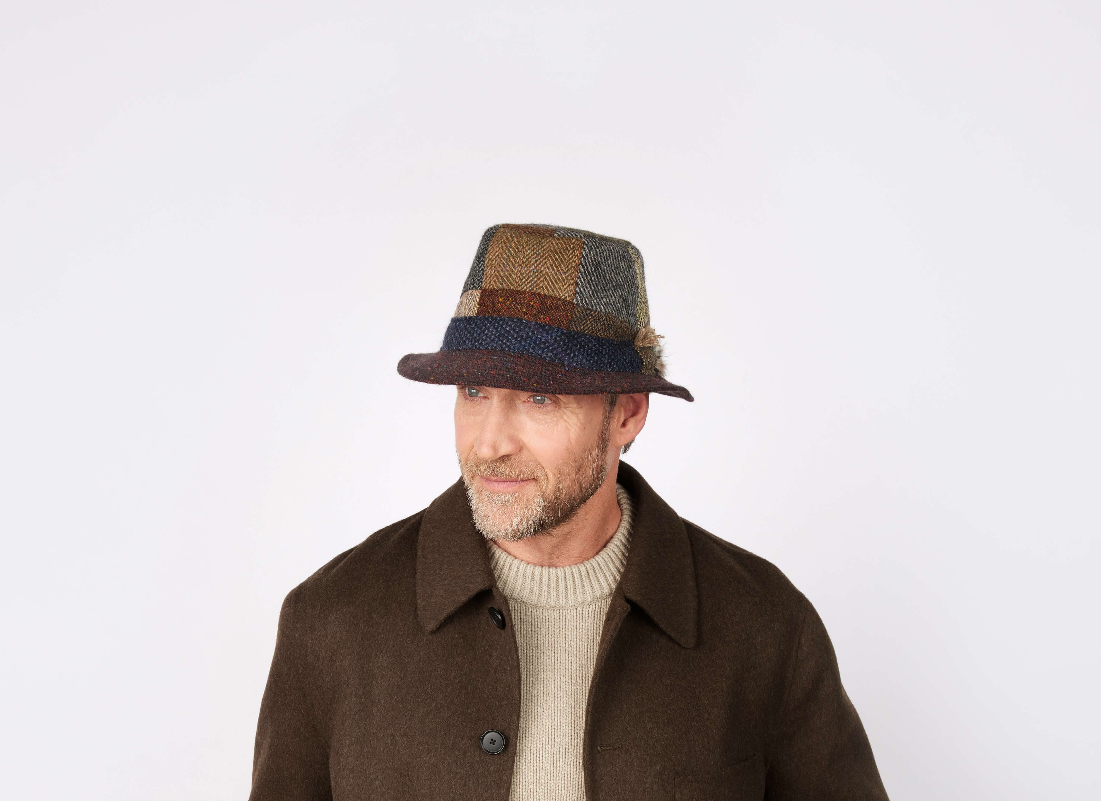 A man with a short beard and light sweater models the Hanna Hats Walking Hat Patchwork Tweed, a brown patchwork hat inspired by Donegal, Ireland, while wearing a brown coat and looking slightly left against a white background.