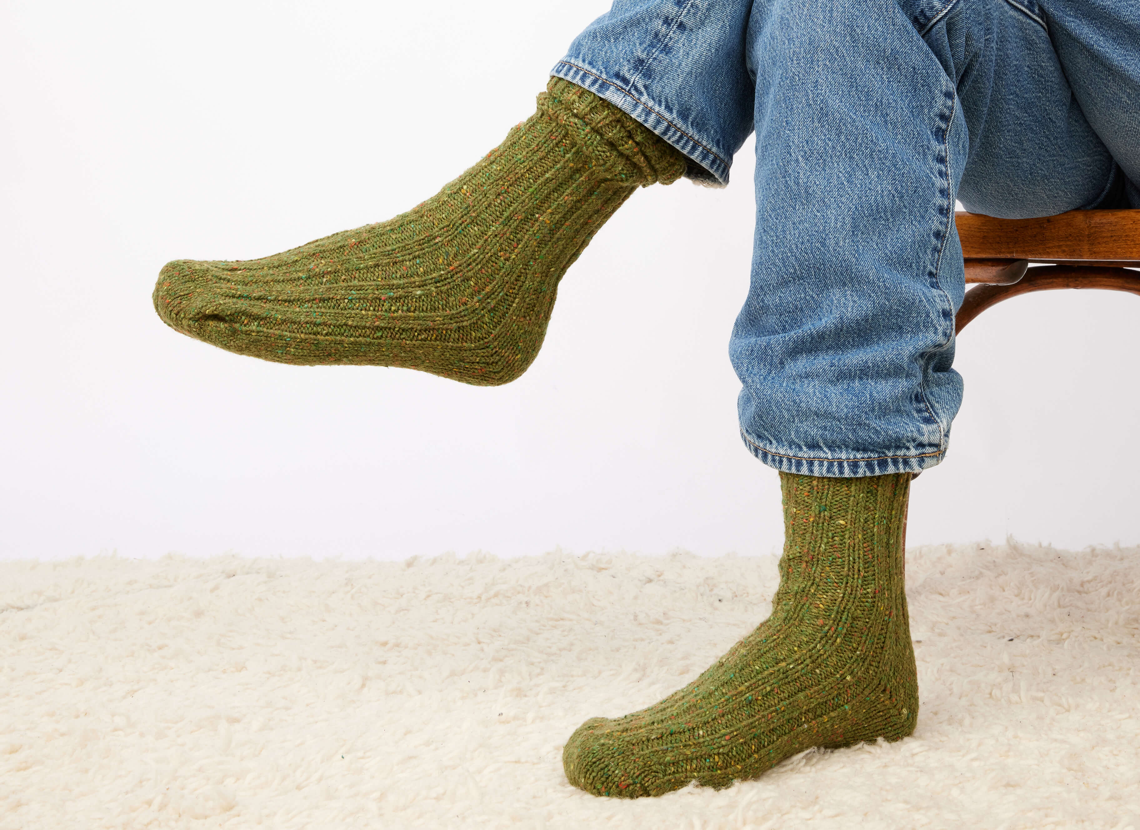 A person wearing blue jeans and Hanna Hats Donegal Socks sits cross-legged on a wooden chair over a light-colored carpet, with only their lower legs and feet visible.