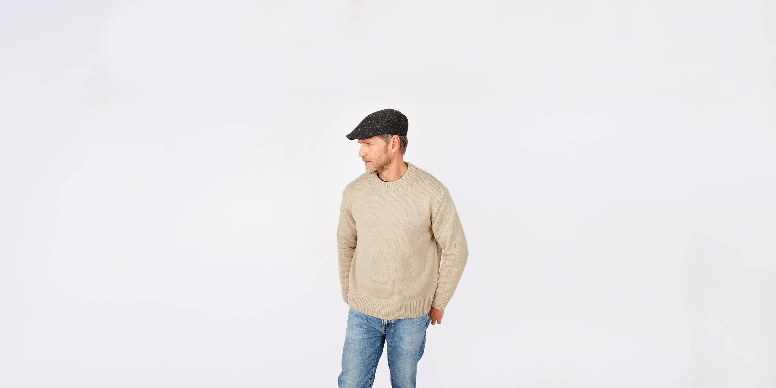 A man wearing a beige sweater, blue jeans, and a dark flat cap stands against a plain white background, looking to his left with his hands in his pockets.