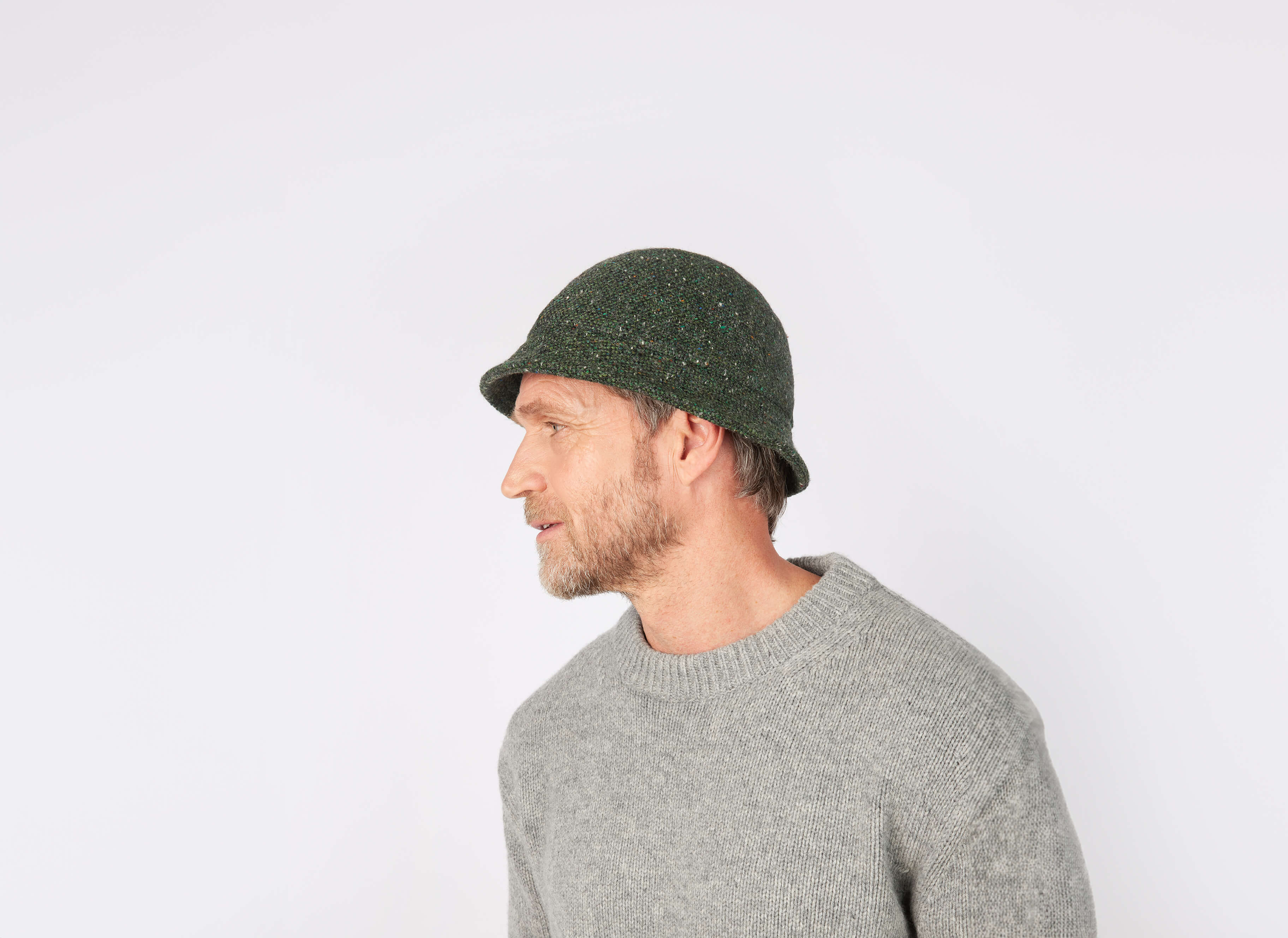A bearded man in profile wears a green Hanna Hats Eske Travel Hat Tweed and a gray sweater against a plain white background.