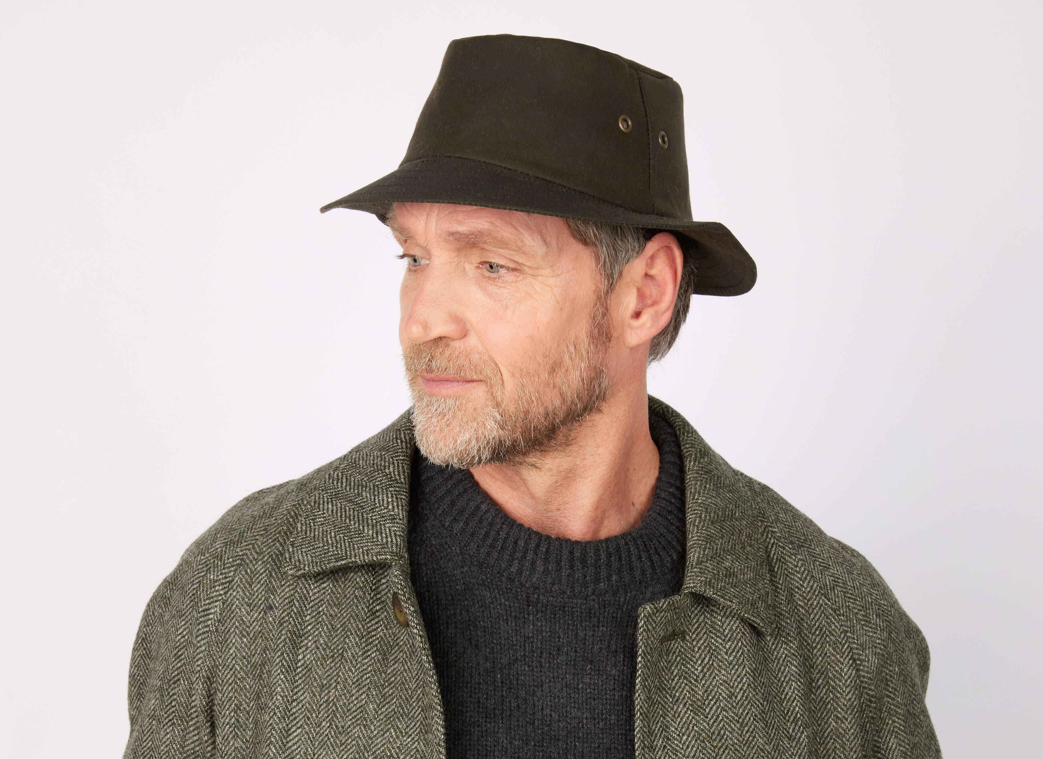 A middle-aged man with a short beard, wearing the Hanna Hats of Donegal Ltd Walking Hat Wax and a grey patterned Irish wax cotton coat, looks slightly to the side against a light background, capturing timeless Donegal style.