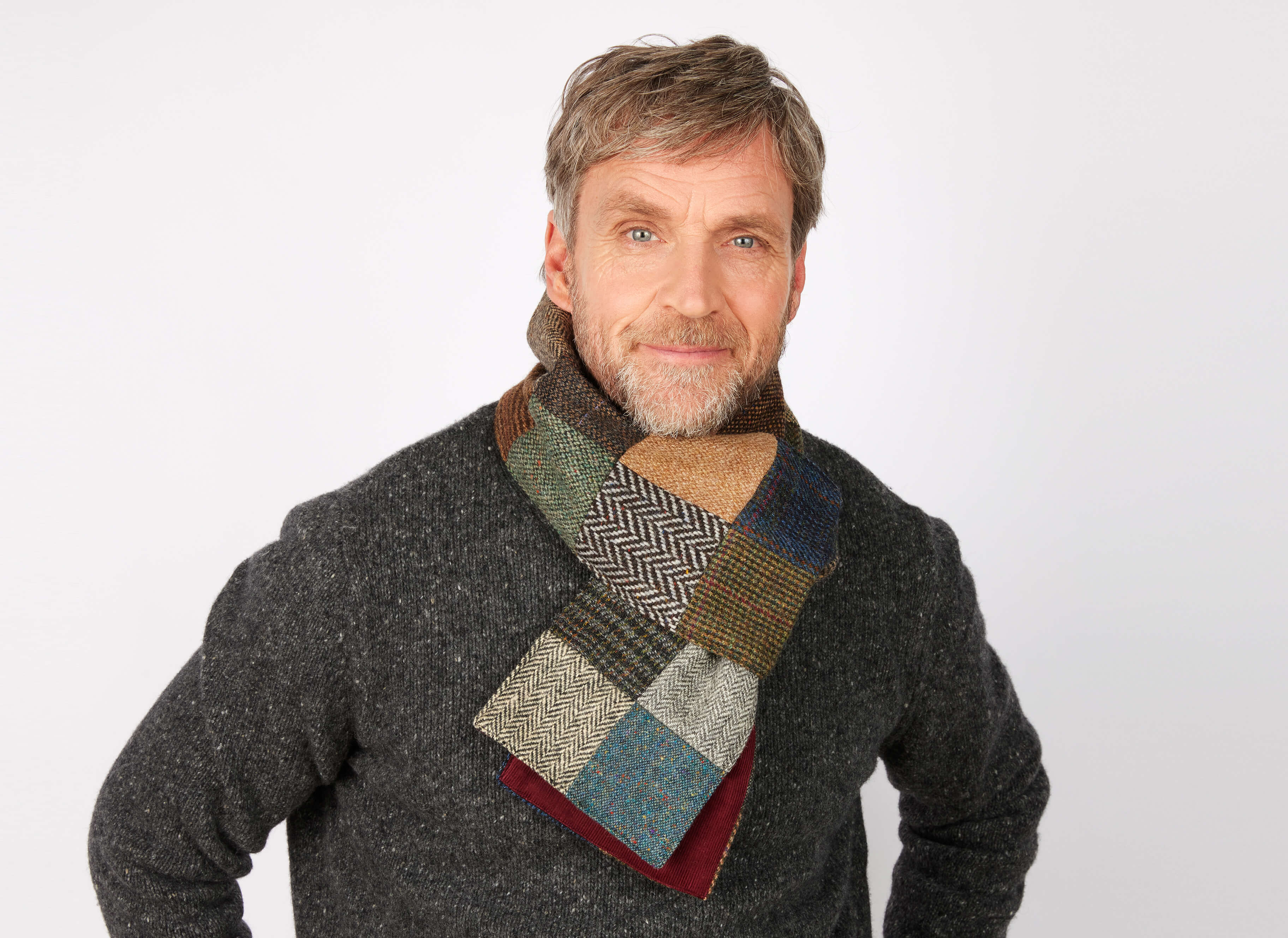 A middle-aged man with gray hair and a beard smiles in a dark sweater, wearing the Hanna Hats Scarf Patchwork Tweed / Corduroy against a plain light background.