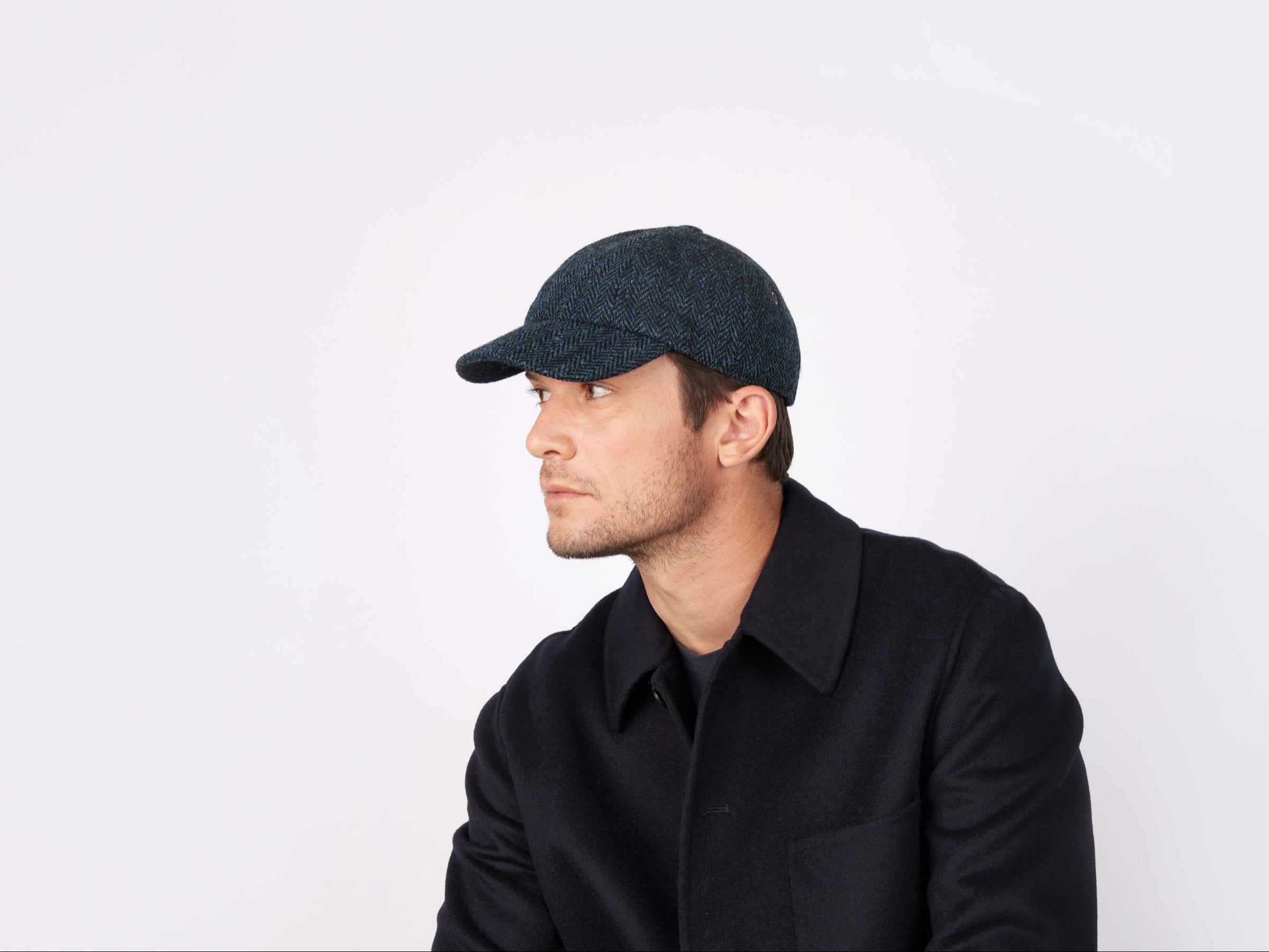 A man in a dark Hanna Hats Baseball Cap Tweed and a dark coat looks to his left against a plain white background.
