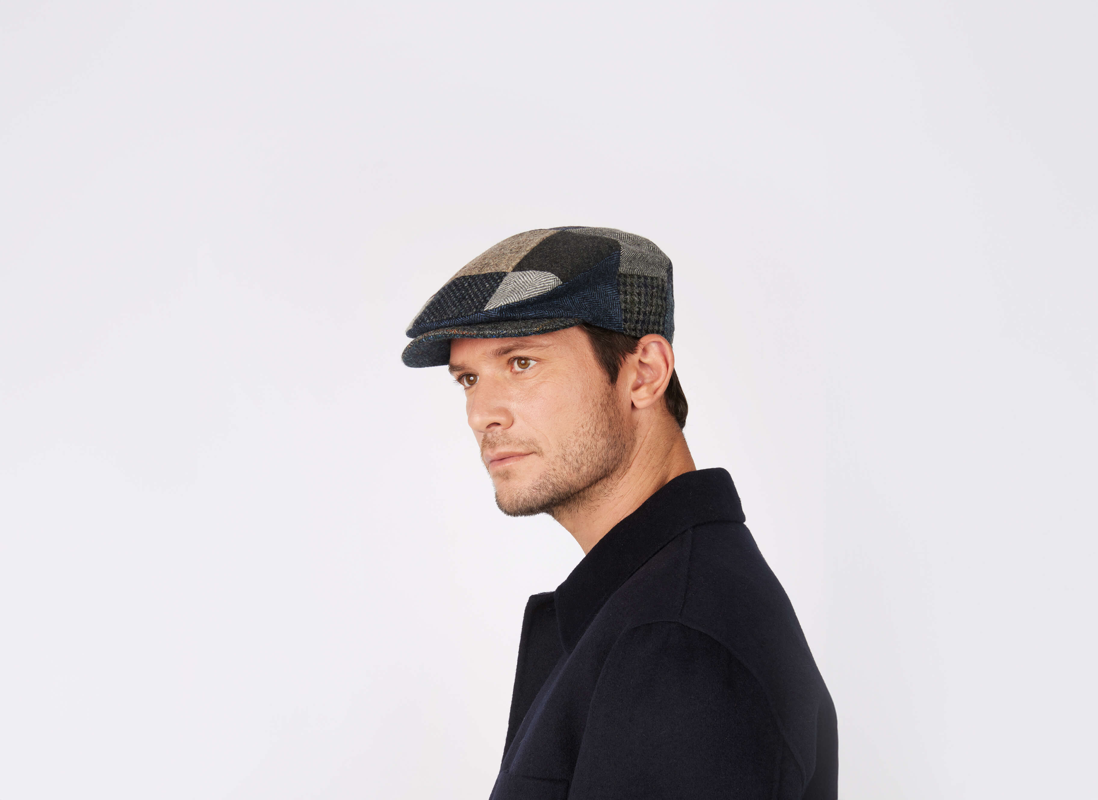 A man in a dark coat wears the Hanna Hats Vintage Cap Patchwork Grey/Blue Tweed, looking to the side against a plain white background.