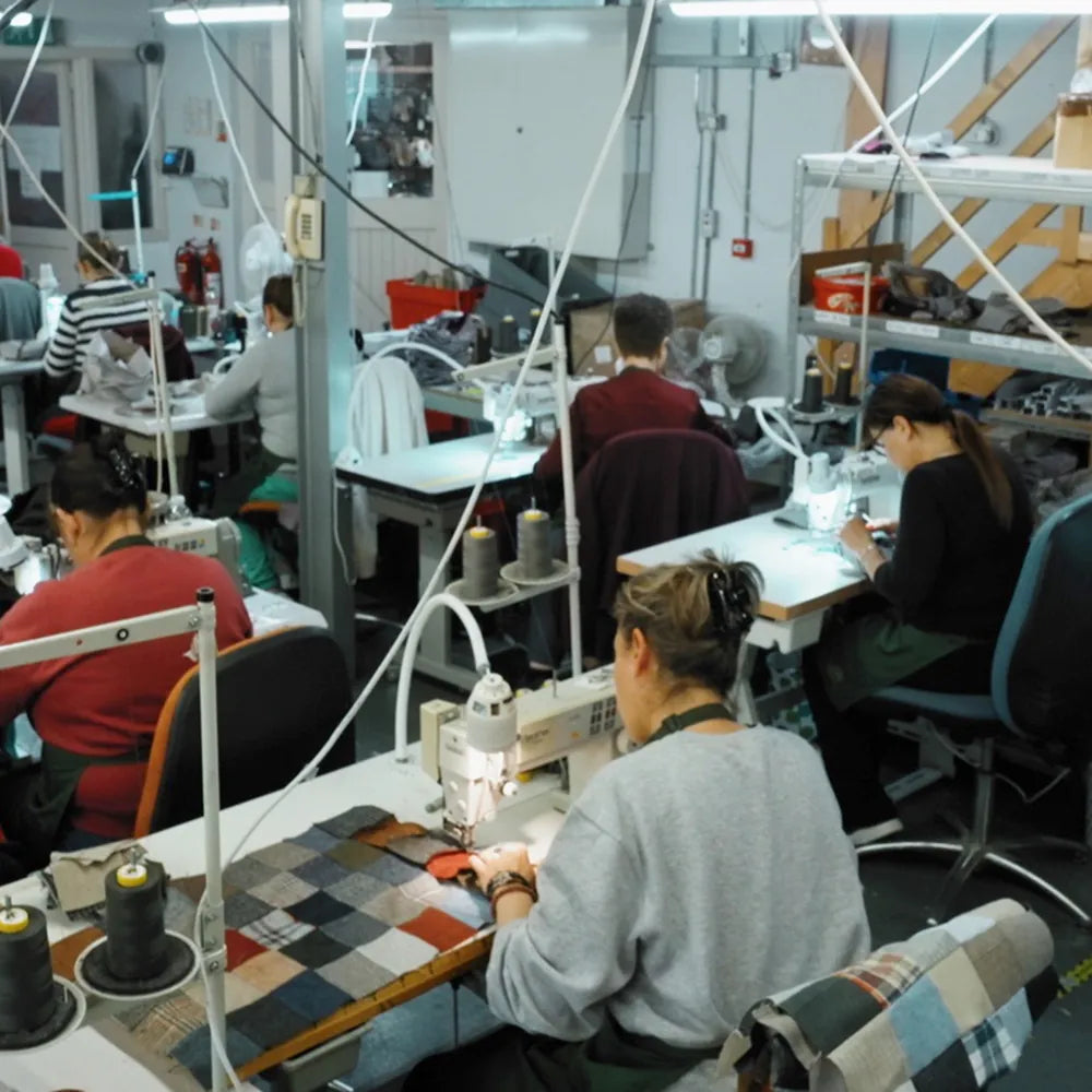 Workers sit at sewing machines in a factory, stitching pieces of fabric. The workspace is busy, with tools, spools of thread, and patchwork materials on tables, and shelves and equipment in the background.